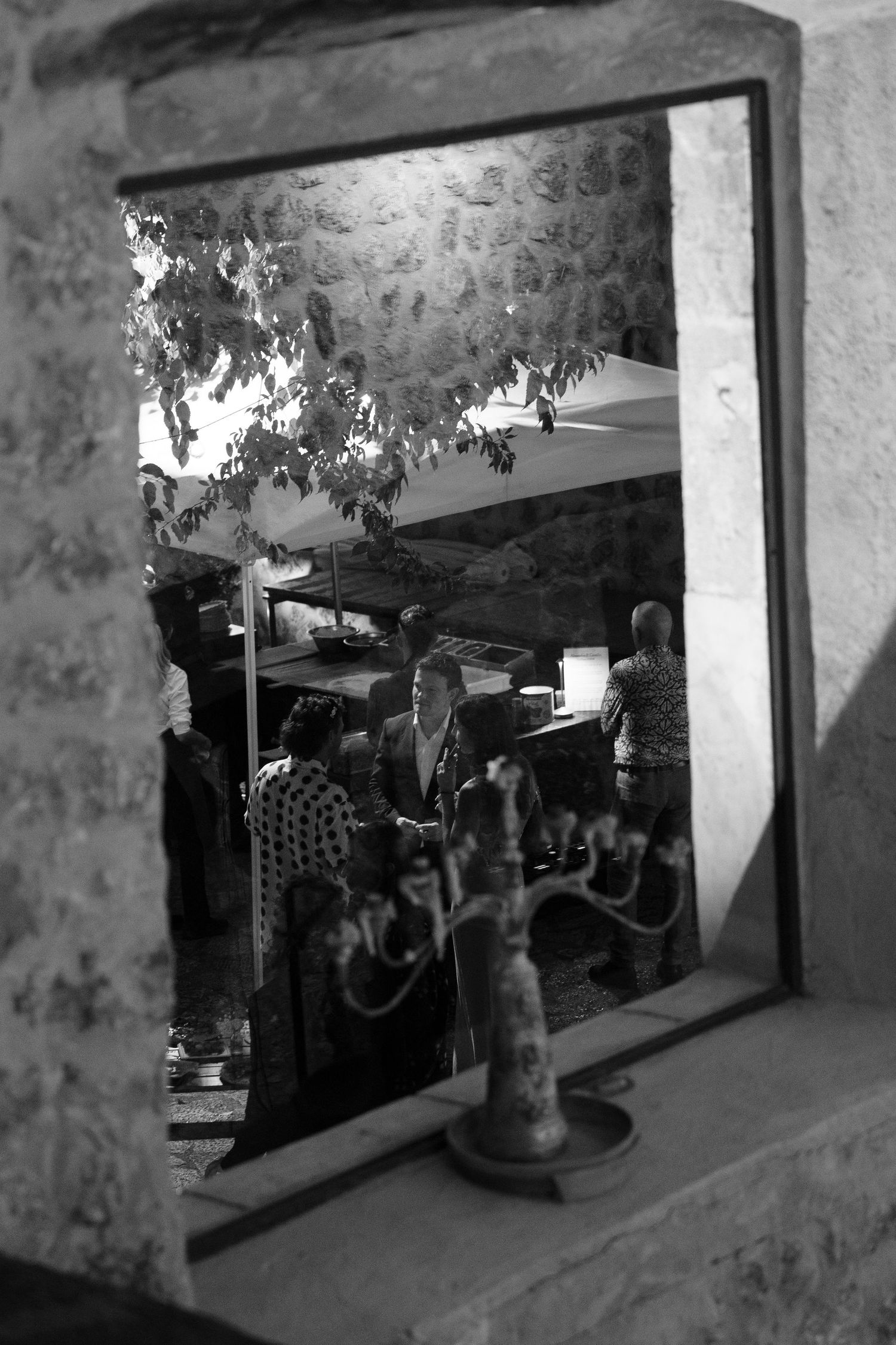 Black and white view through a stone window frame of guests mingling at an outdoor evening gathering beneath trees and canopies.