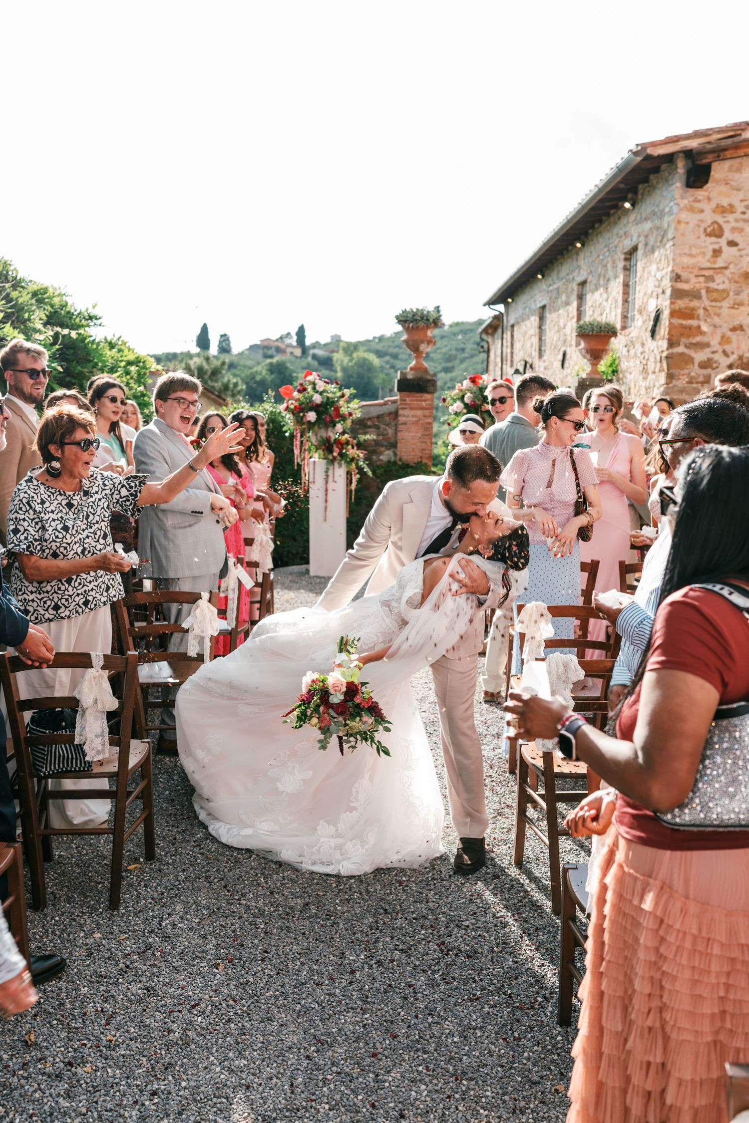 Groom dips and kisses his bride in a white gown as smiling guests cheer along a gravel aisle at a rustic Tuscan stone villa wedding.