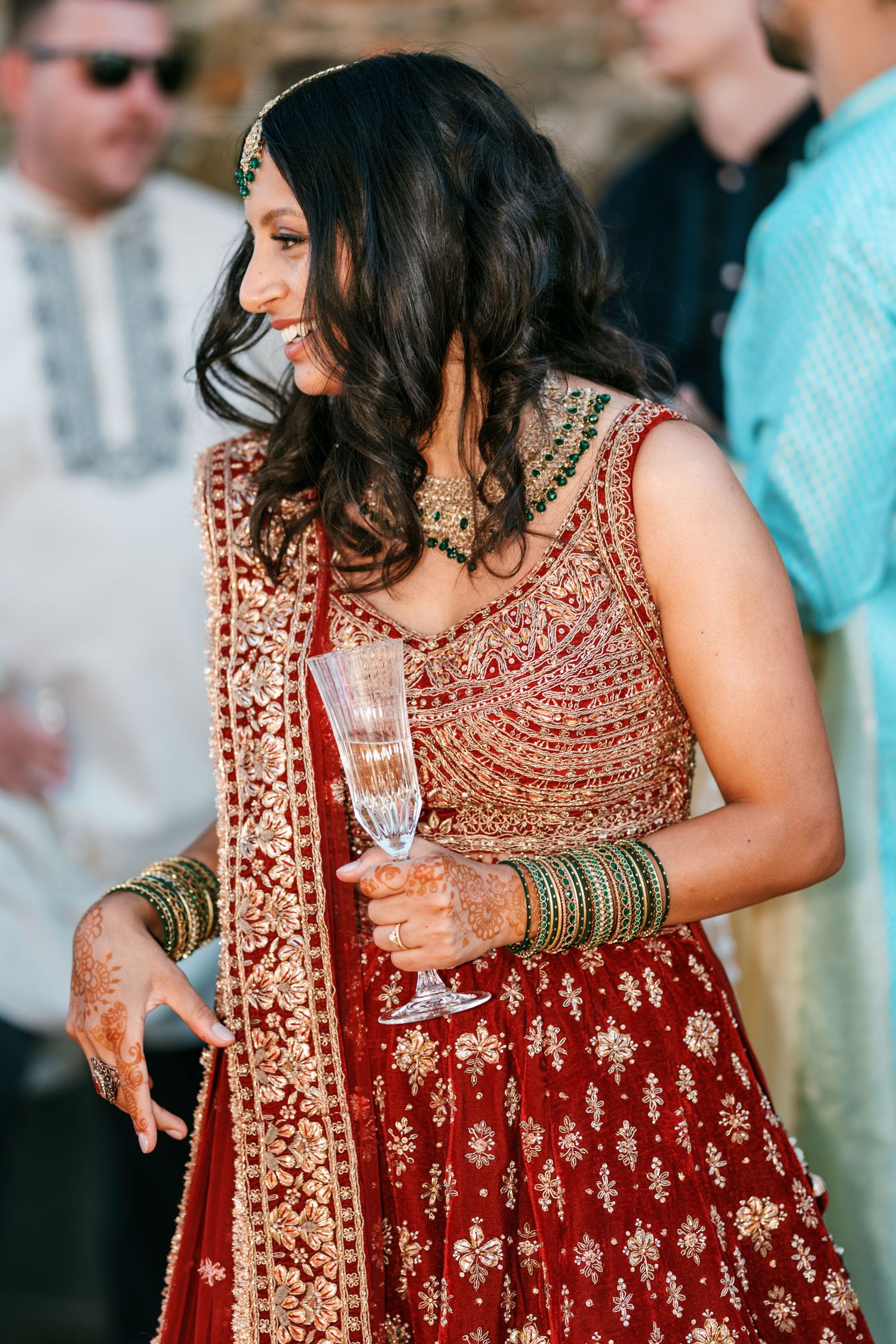 Smiling bride with henna-decorated hands and green bangles holds a champagne flute in a red and gold embroidered lehenga.