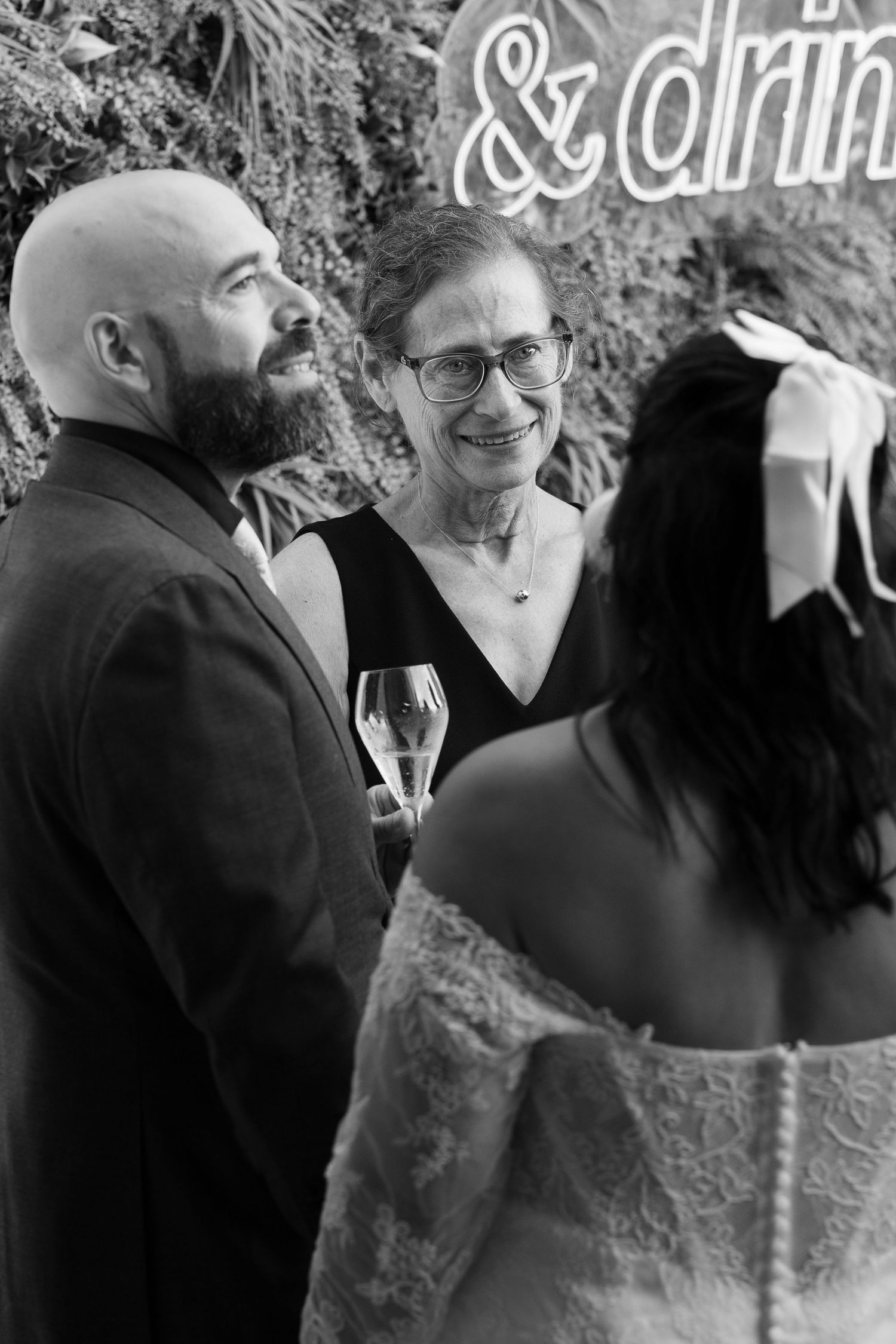 Black and white photo of wedding guests smiling and holding champagne near a neon sign, with a bride in a lace gown in the foreground.