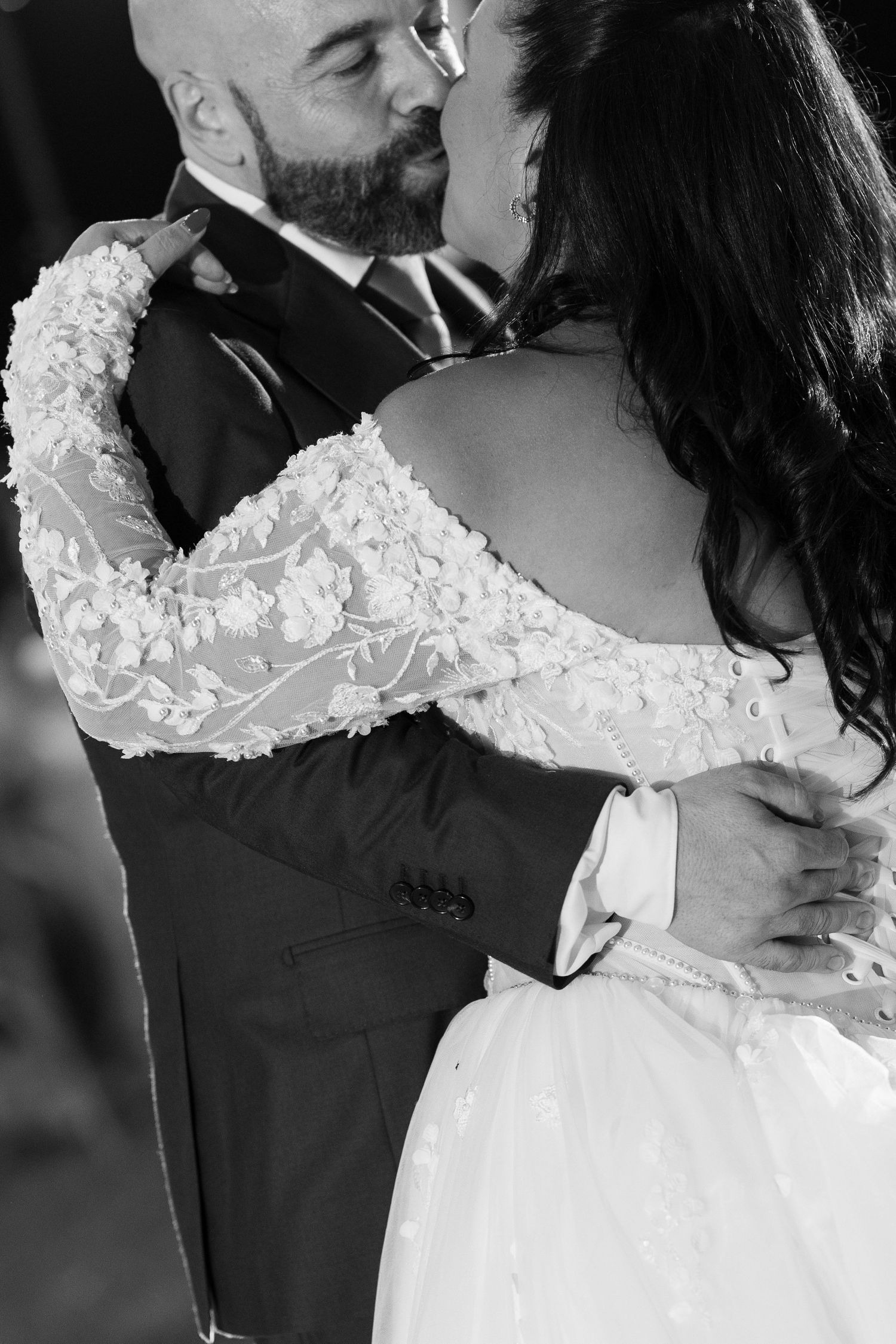 Black and white wedding photo of a bearded groom in a suit kissing a bride in an off-shoulder lace gown with long sleeves.