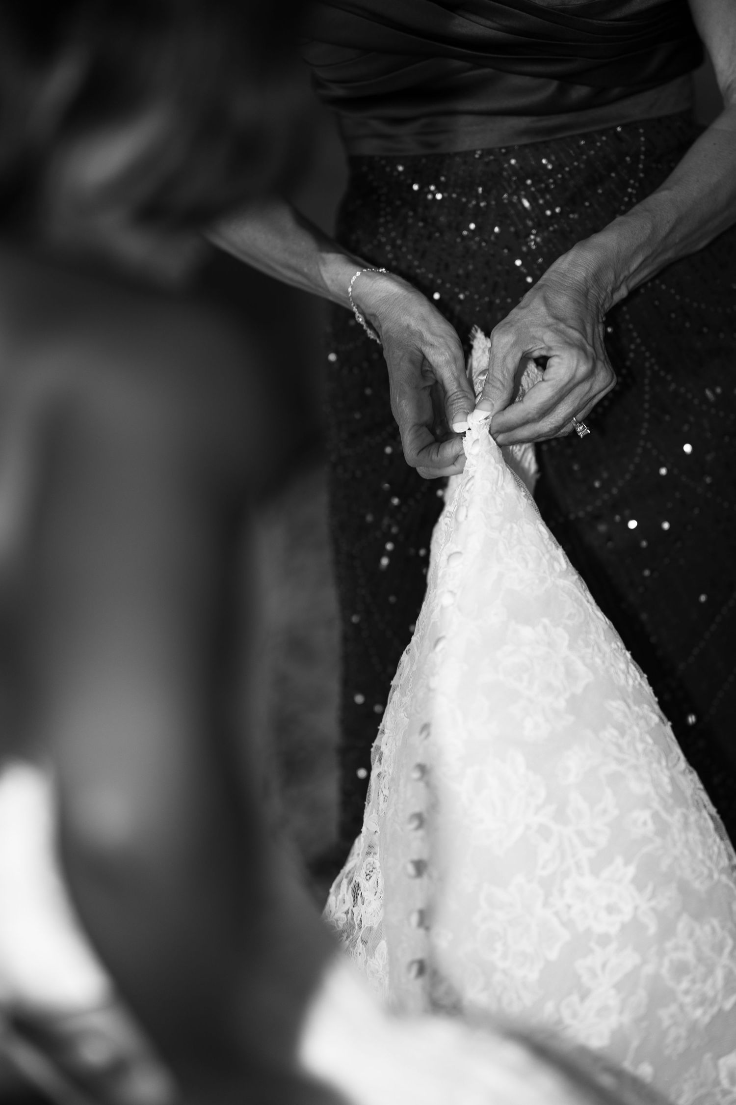 Black and white photo of a mother in a sequined dress fastening the lace buttons on a bride's wedding gown.