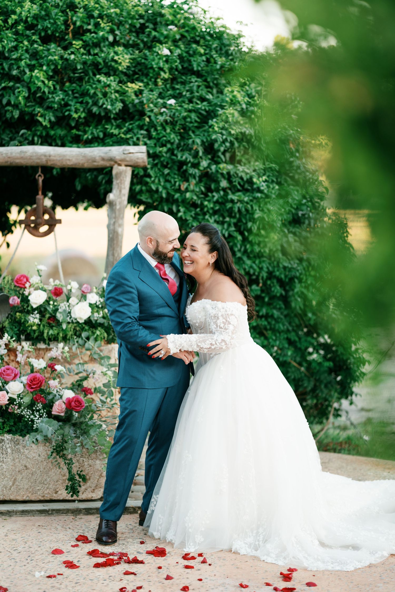Smiling bride in an off-shoulder lace wedding gown embraces her groom in a teal suit amid scattered rose petals and a lush garden.