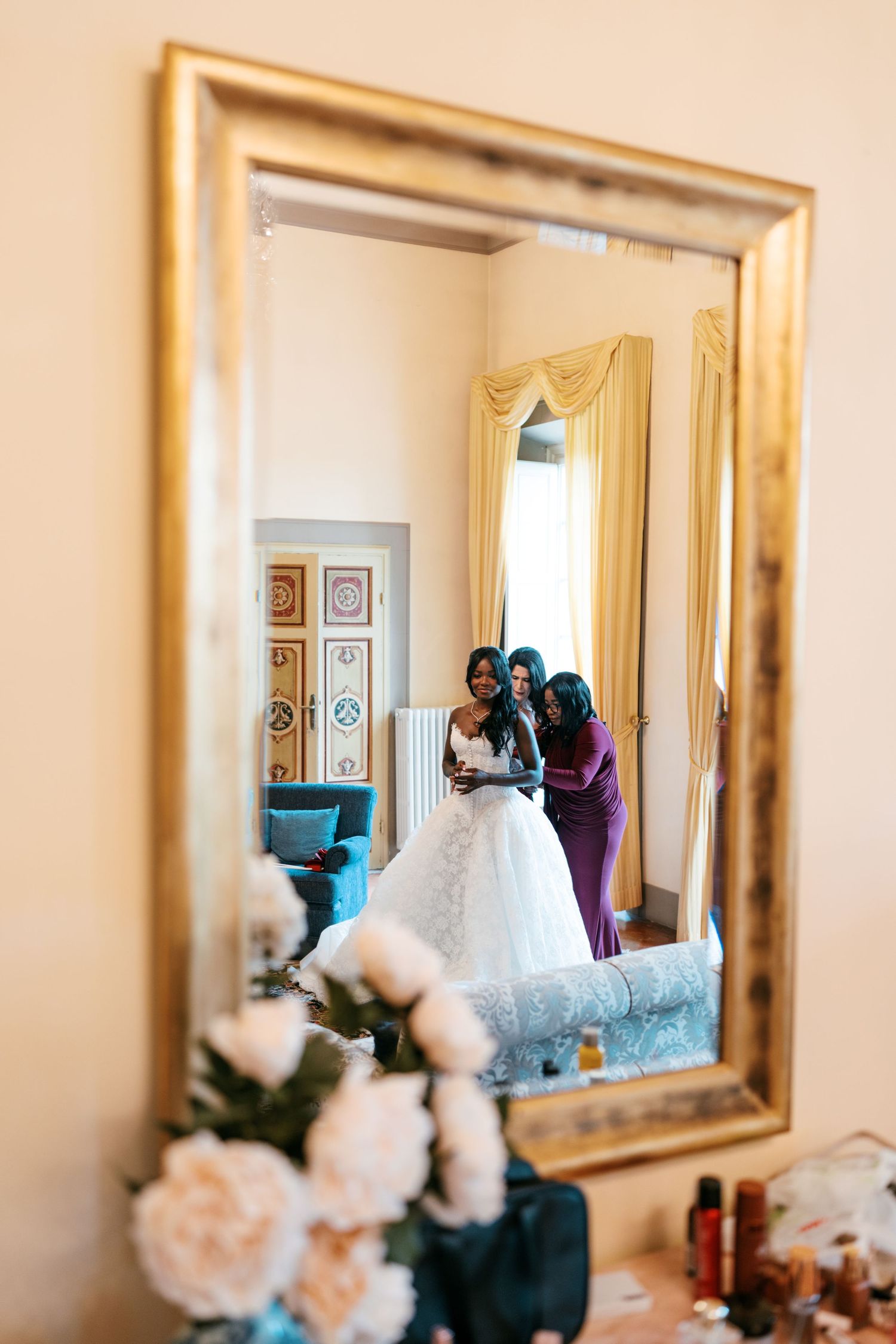 Bride in a white lace wedding gown getting ready with two women, reflected in a gold-framed mirror beside white peony flowers.