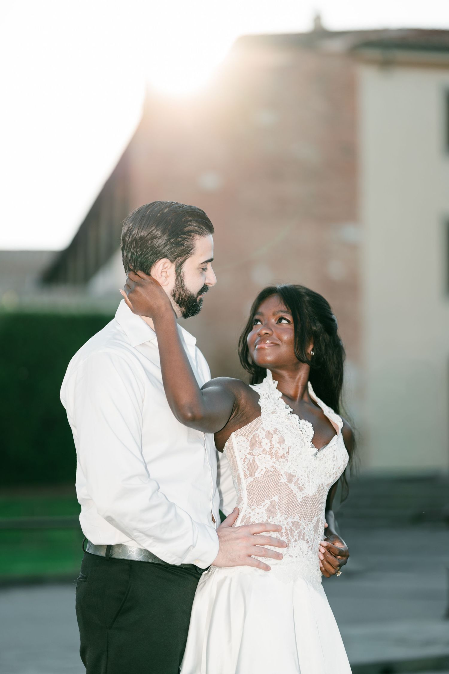 Romantic couple in a white lace dress and white shirt embrace outdoors at golden hour, smiling at each other in an engagement photo.