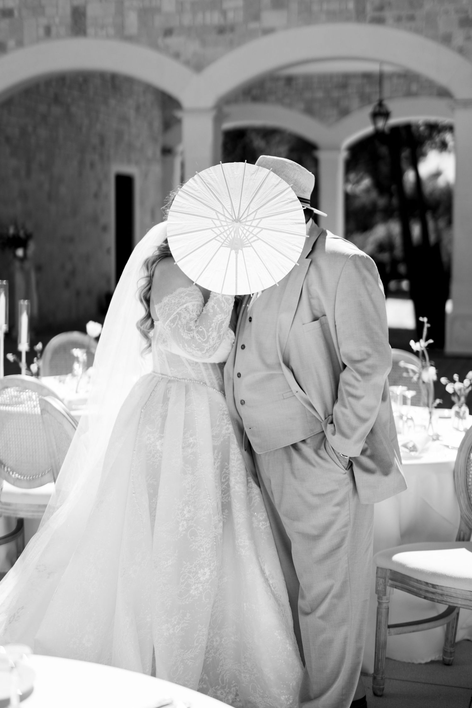 Black and white photo of bride and groom kissing behind a white parasol at elegant wedding reception in Mallorca.