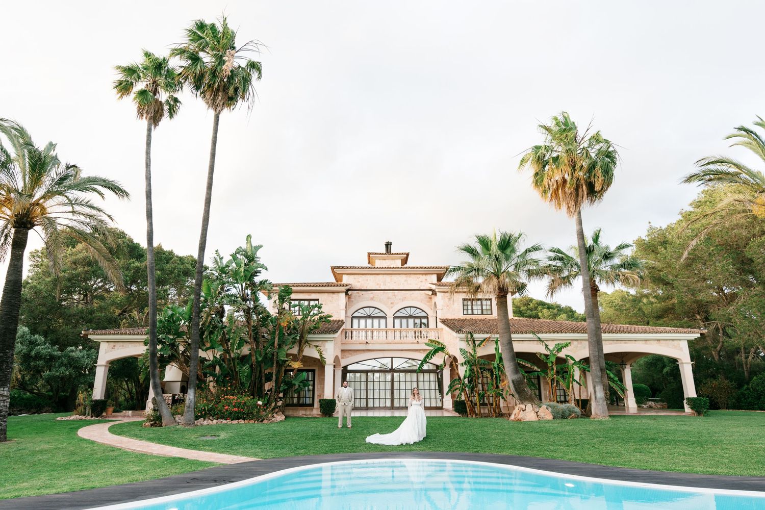 Bride in white gown poses by pool at luxury Mediterranean villa with tall palm trees and lush gardens.
