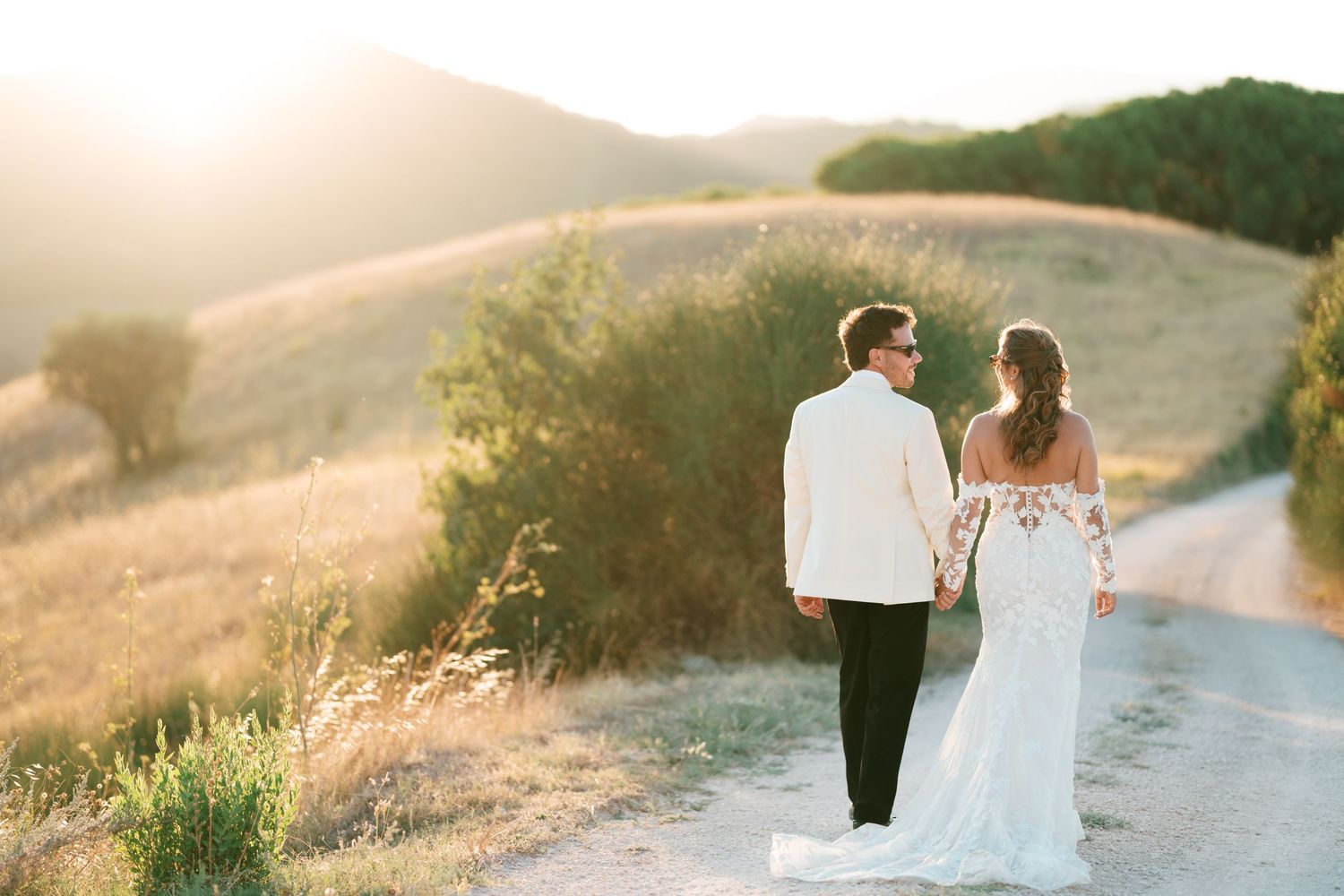 Bride and groom holding hands walking on a sunlit path with golden hills in the background.