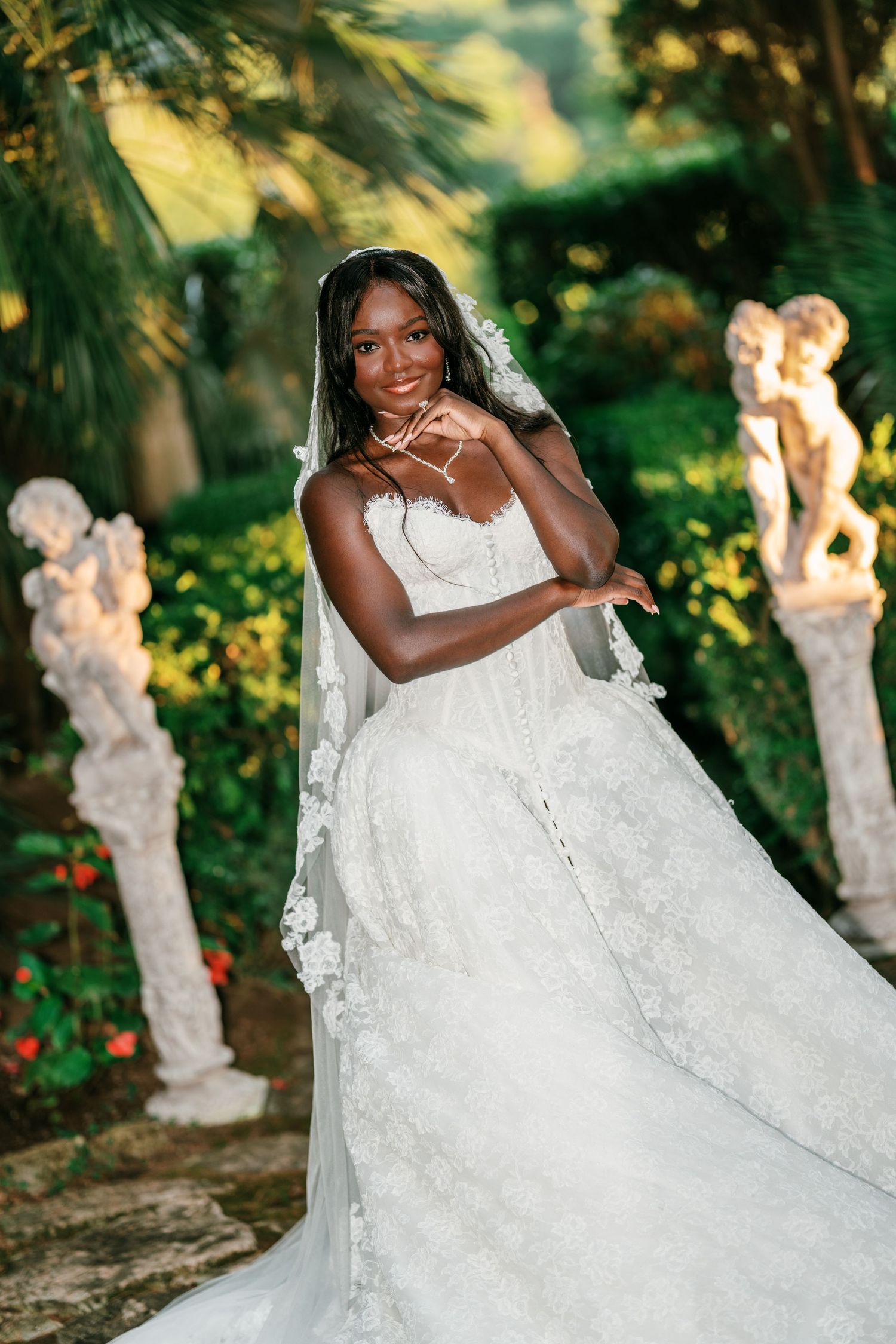 Smiling bride in a lace wedding gown and veil poses gracefully near cherub statues in a sunlit garden.