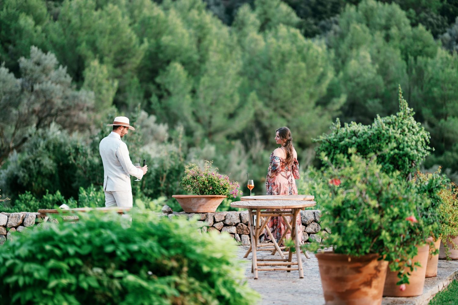 Man in a straw hat and woman in a floral dress chat beside a wooden table surrounded by terracotta pots in a lush garden.