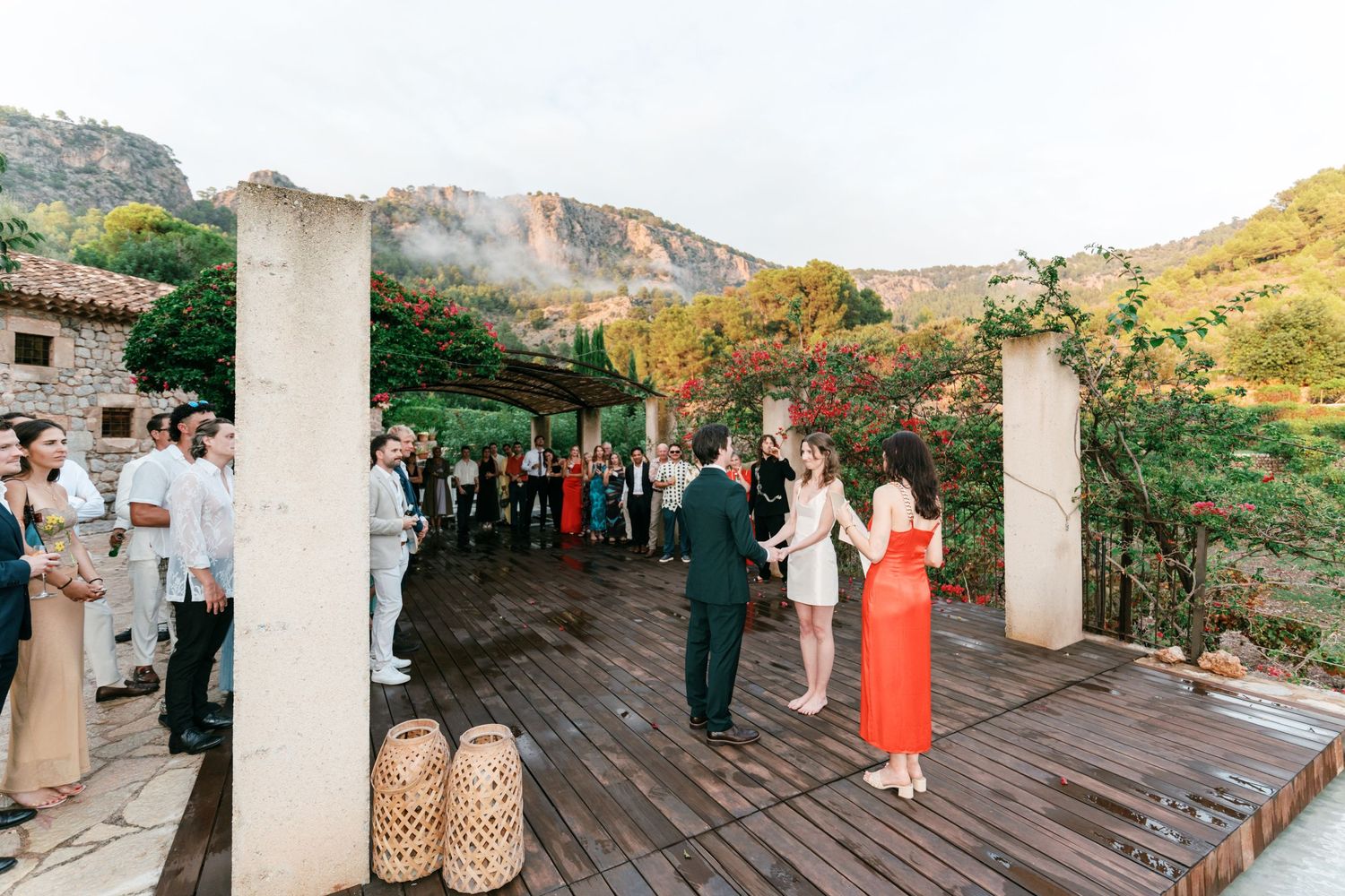 Wedding guests gather on a wooden terrace with a bride in a red dress and groom in a suit, set against scenic mountains.