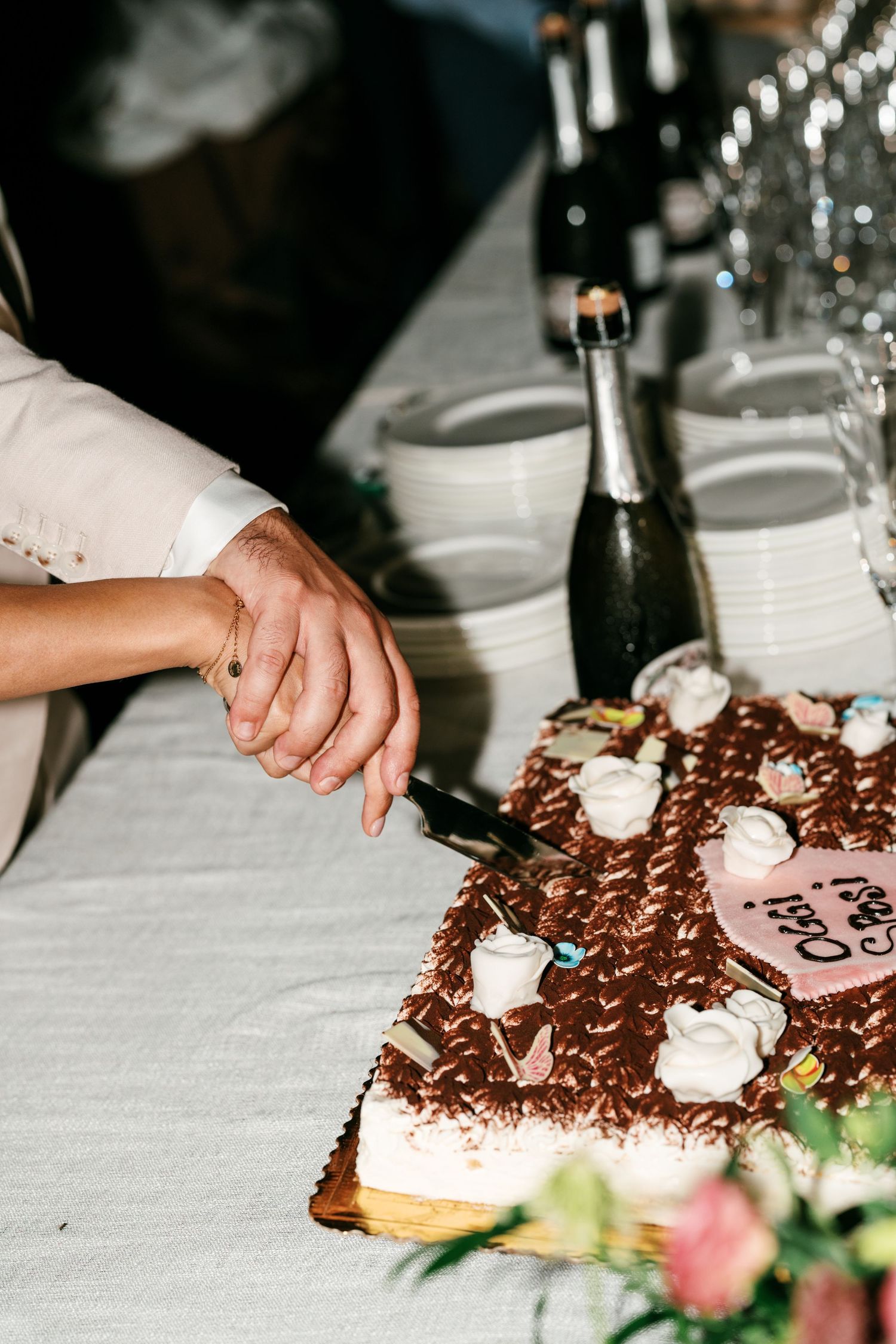 Couple's hands cutting a chocolate wedding cake decorated with white sugar roses, with champagne bottles and stacked plates behind.