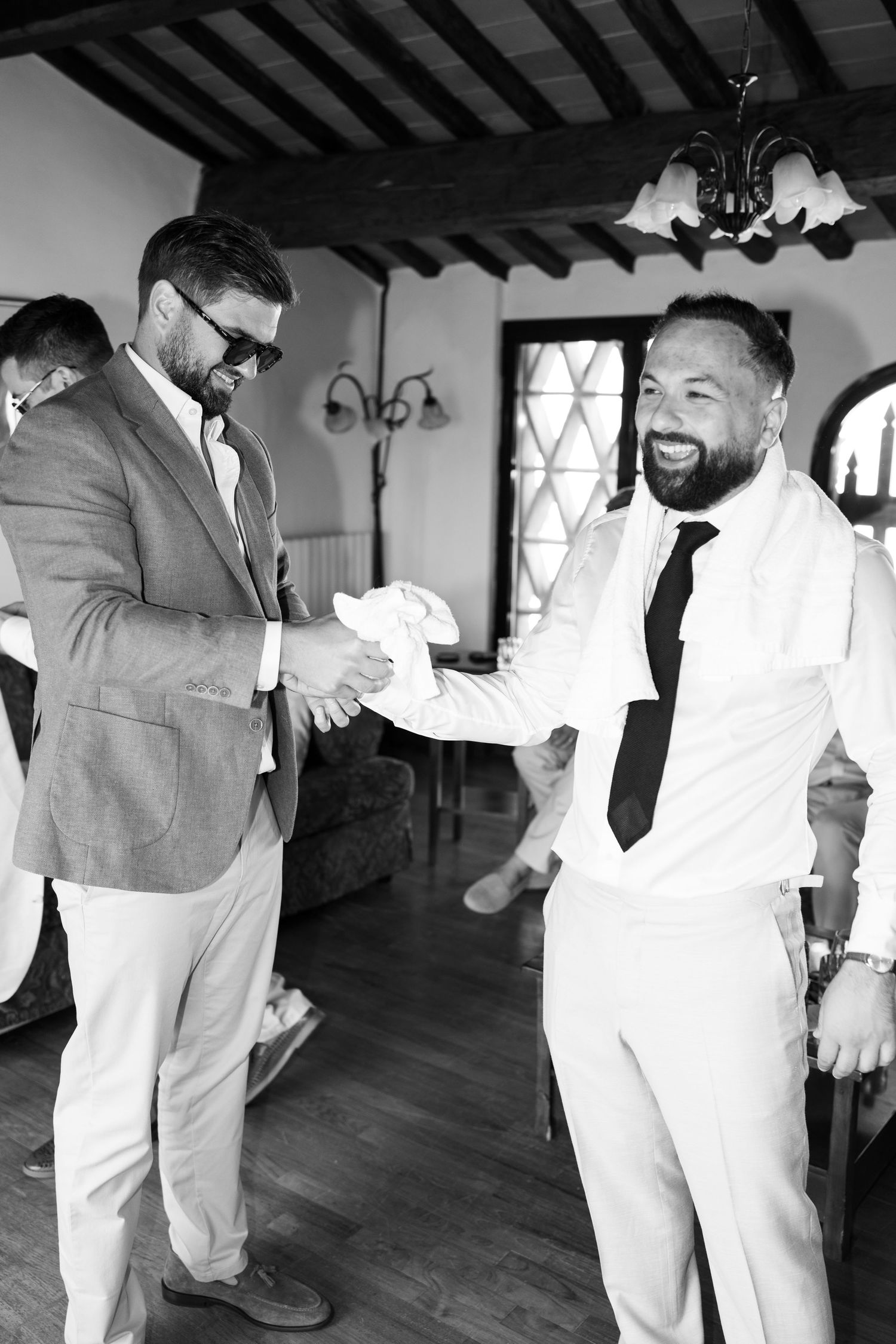 Two smiling groomsmen in a rustic room shake hands in black and white, one in sunglasses and blazer, the other in a vest and tie.