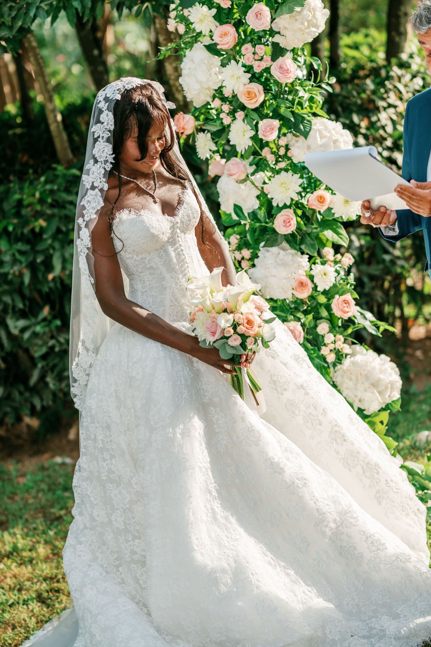 Bride in a strapless lace wedding gown and lace-trimmed veil holds a pink bouquet beside a floral arch at an outdoor garden ceremony.