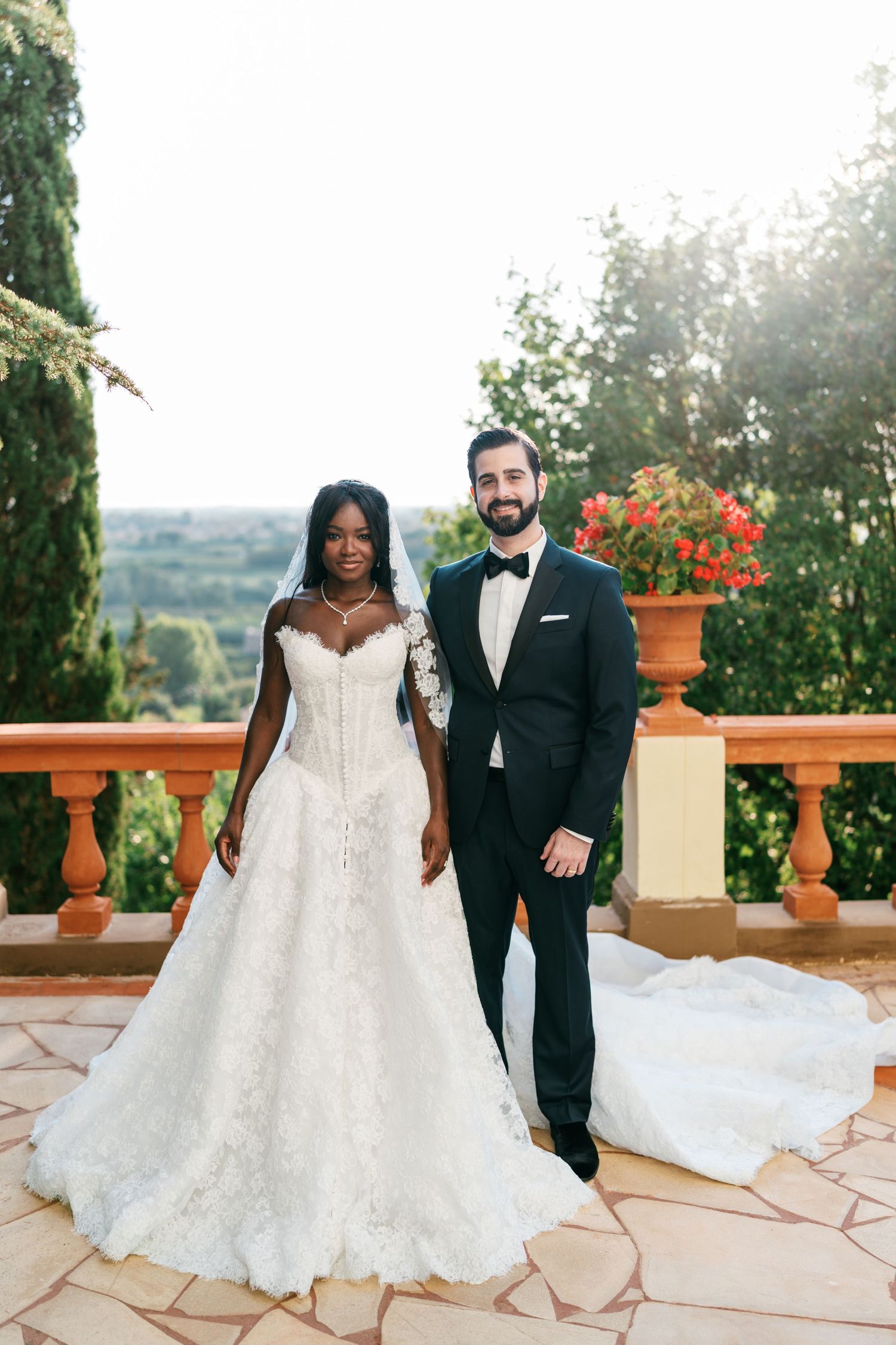 Bride in a lace off-the-shoulder gown and groom in a black tuxedo pose on a terracotta terrace with a scenic hillside view.