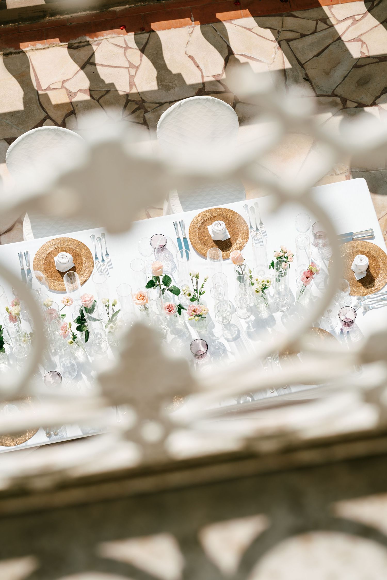 Elegant outdoor wedding table with woven chargers, pink floral centerpieces and glassware, viewed through an ornate white iron railing.