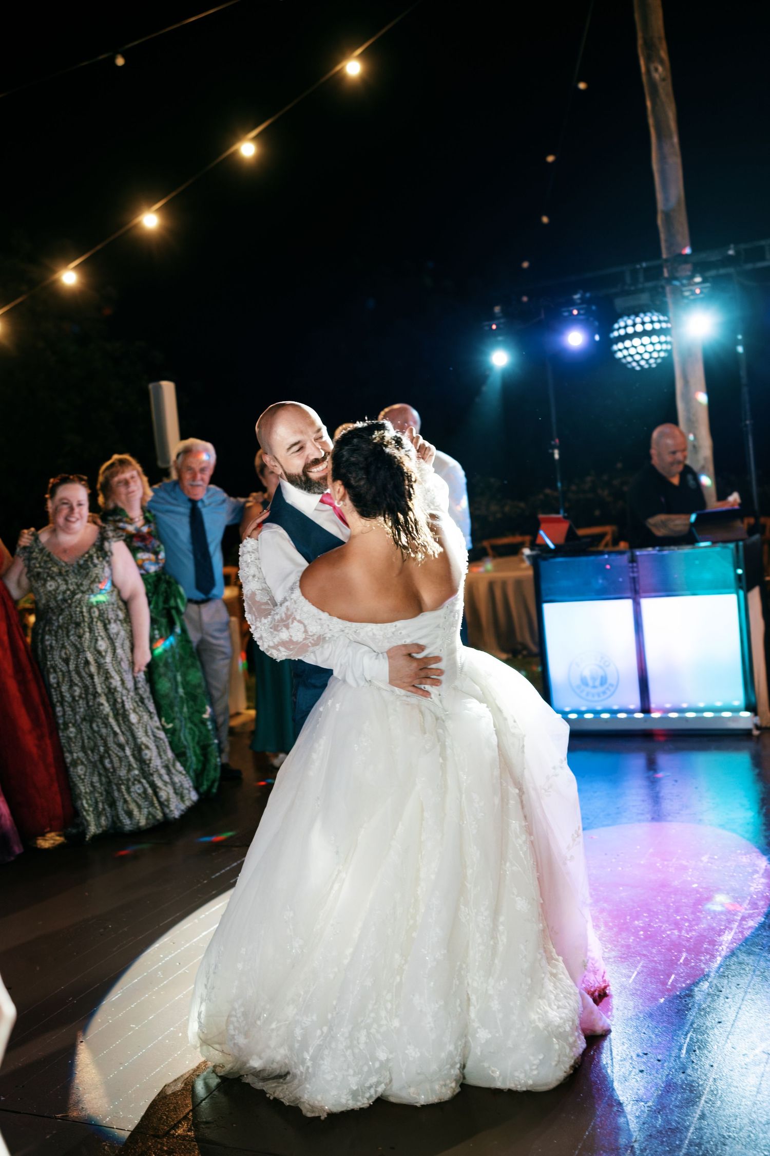 Bride and groom share a romantic first dance as wedding guests watch beneath string lights and a mirror ball at night.