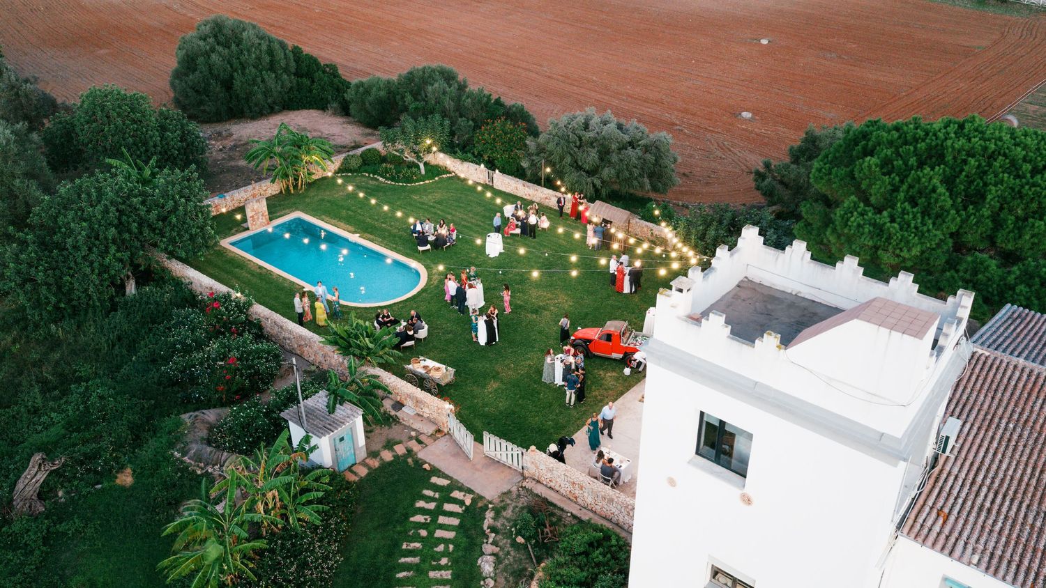 Aerial view of an outdoor garden party beside a swimming pool at a Mediterranean villa with string lights, guests, and surrounding trees.