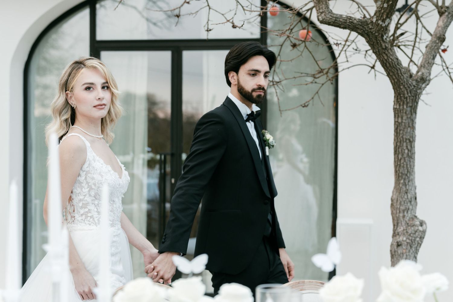 Bride and groom holding hands at a floral-decorated wedding table near an arched white building. Mallorca wedding photographer, filmmaker