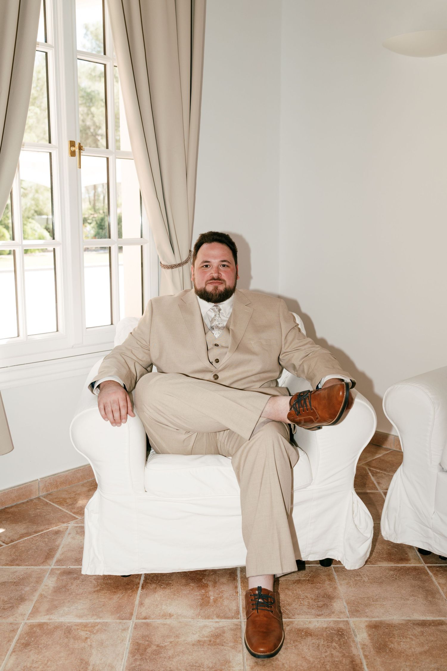 A well-dressed groom in a beige suit sits relaxed in a white armchair near a sunlit window with elegant curtains in Mallorca.