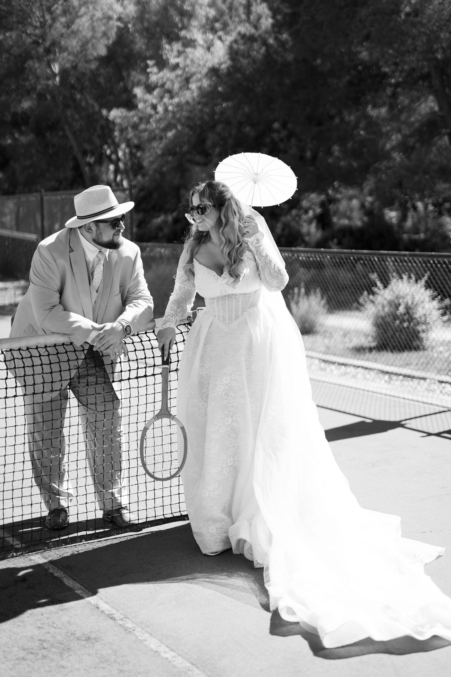Black and white photo of bride and groom posing at tennis net with racket and parasol outdoors.