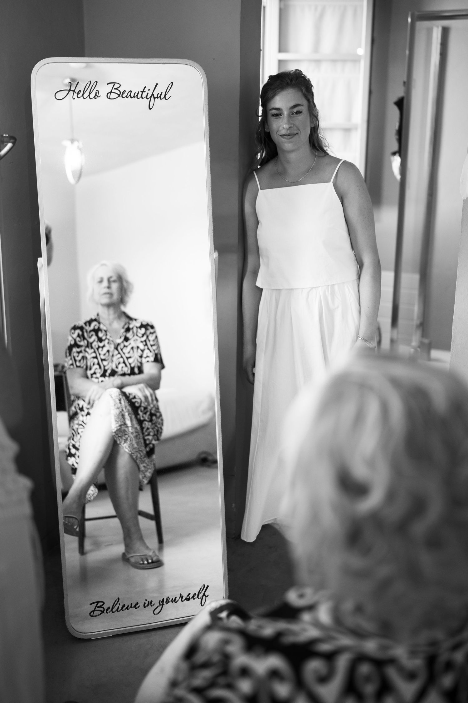 Black and white photo of bride in white dress standing before mirror with older woman seated, mirror reads Hello Beautiful.