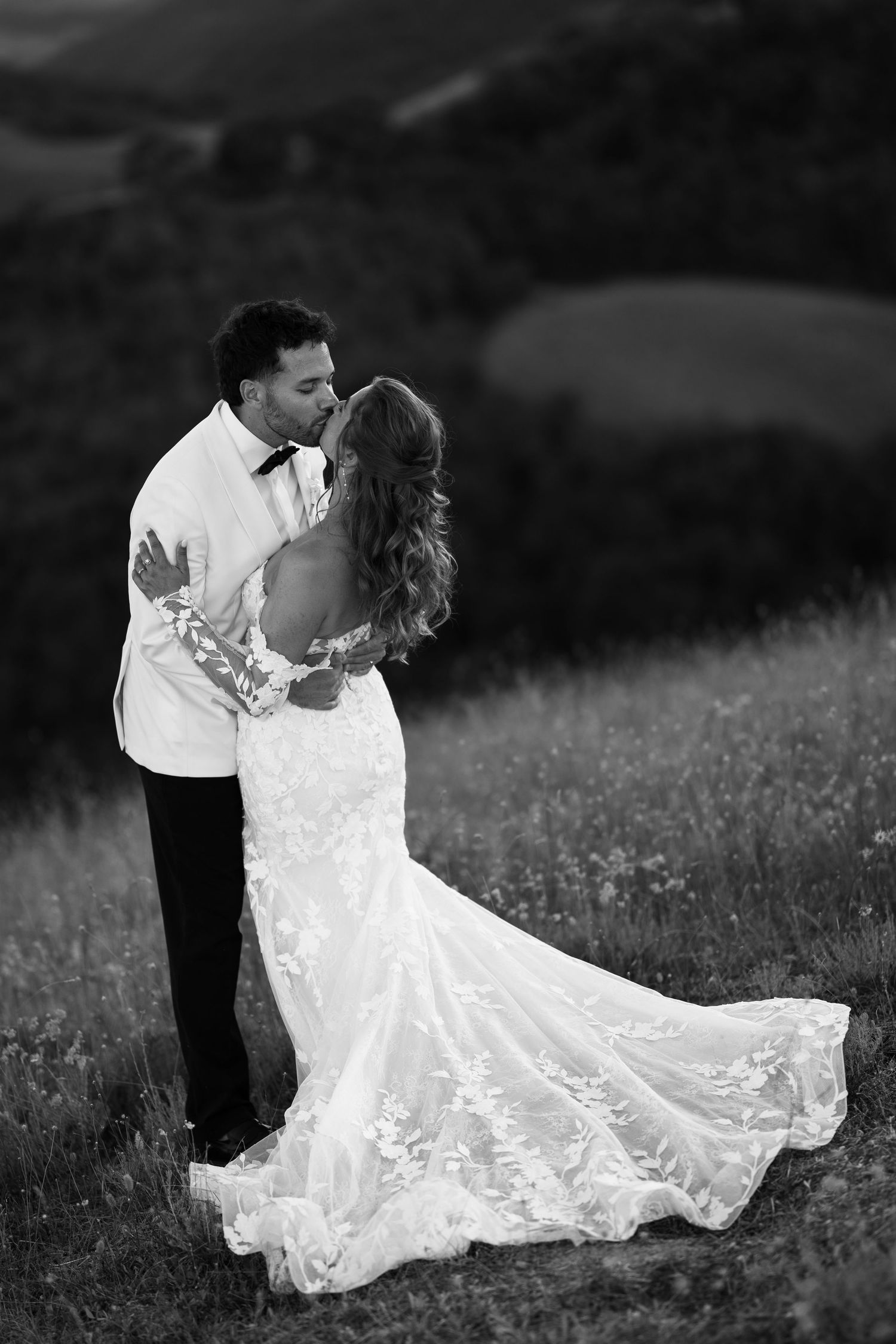 Black and white photo of a bride and groom sharing a romantic kiss in a scenic outdoor field on their wedding day.