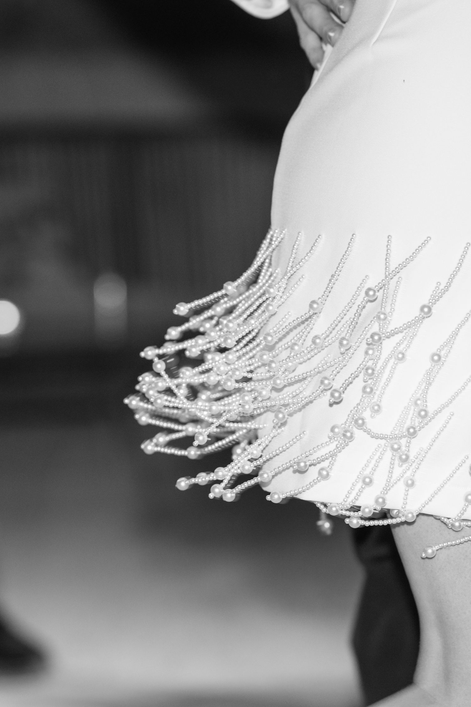 Black and white close-up of elegant beaded fringe dress sleeve with pearl embellishments at a formal event.