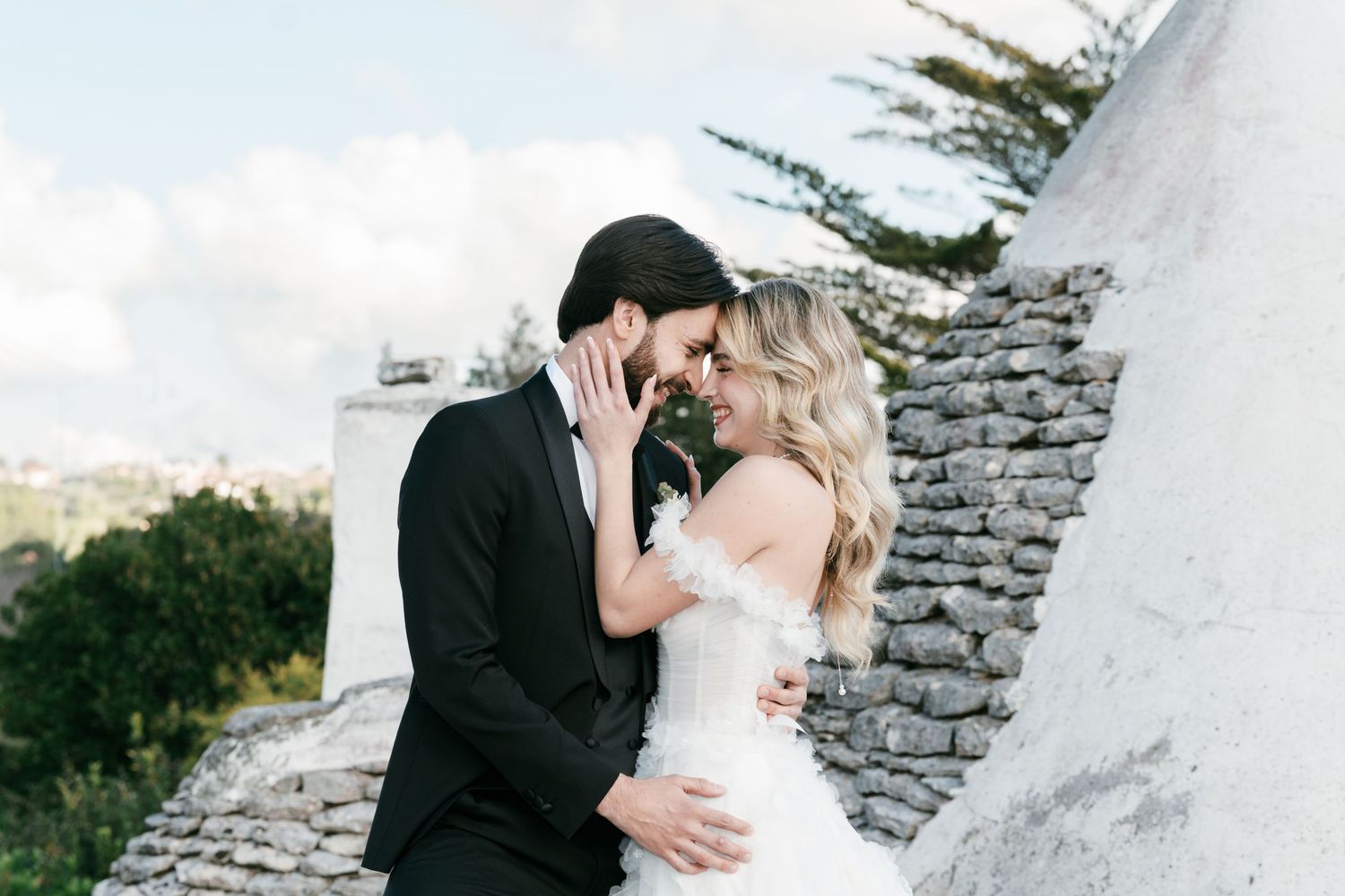 Bride and groom share a romantic forehead-to-forehead moment beside a rustic stone trulli. Puglia wedding photographer, film