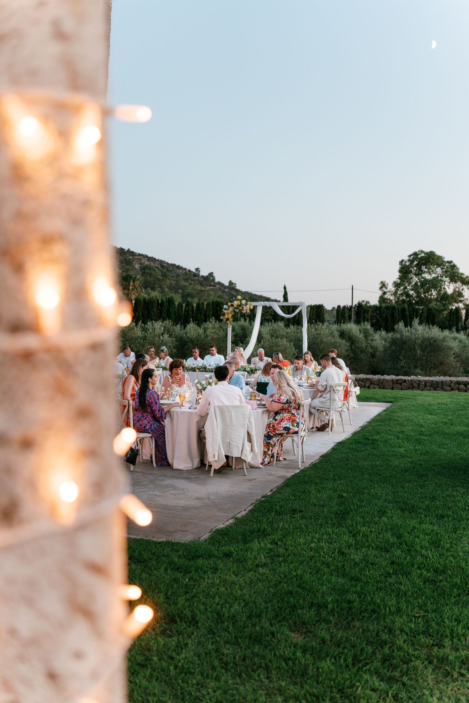 Elegant outdoor wedding reception dinner at dusk with string lights and lush green garden backdrop. Tuscany wedding photographer, filmmaker