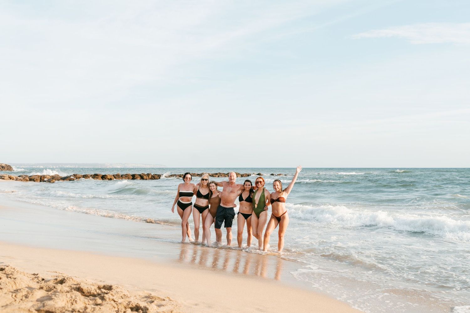 Group of friends in swimwear posing together on a  sandy beach with ocean waves in the background. Mallorca wedding filmmaker, videographer