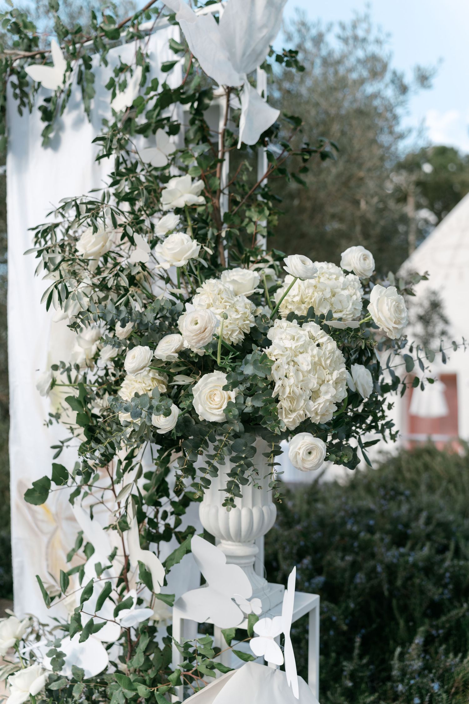 White roses and hydrangeas in ornate vase at outdoor wedding ceremony with greenery backdrop.