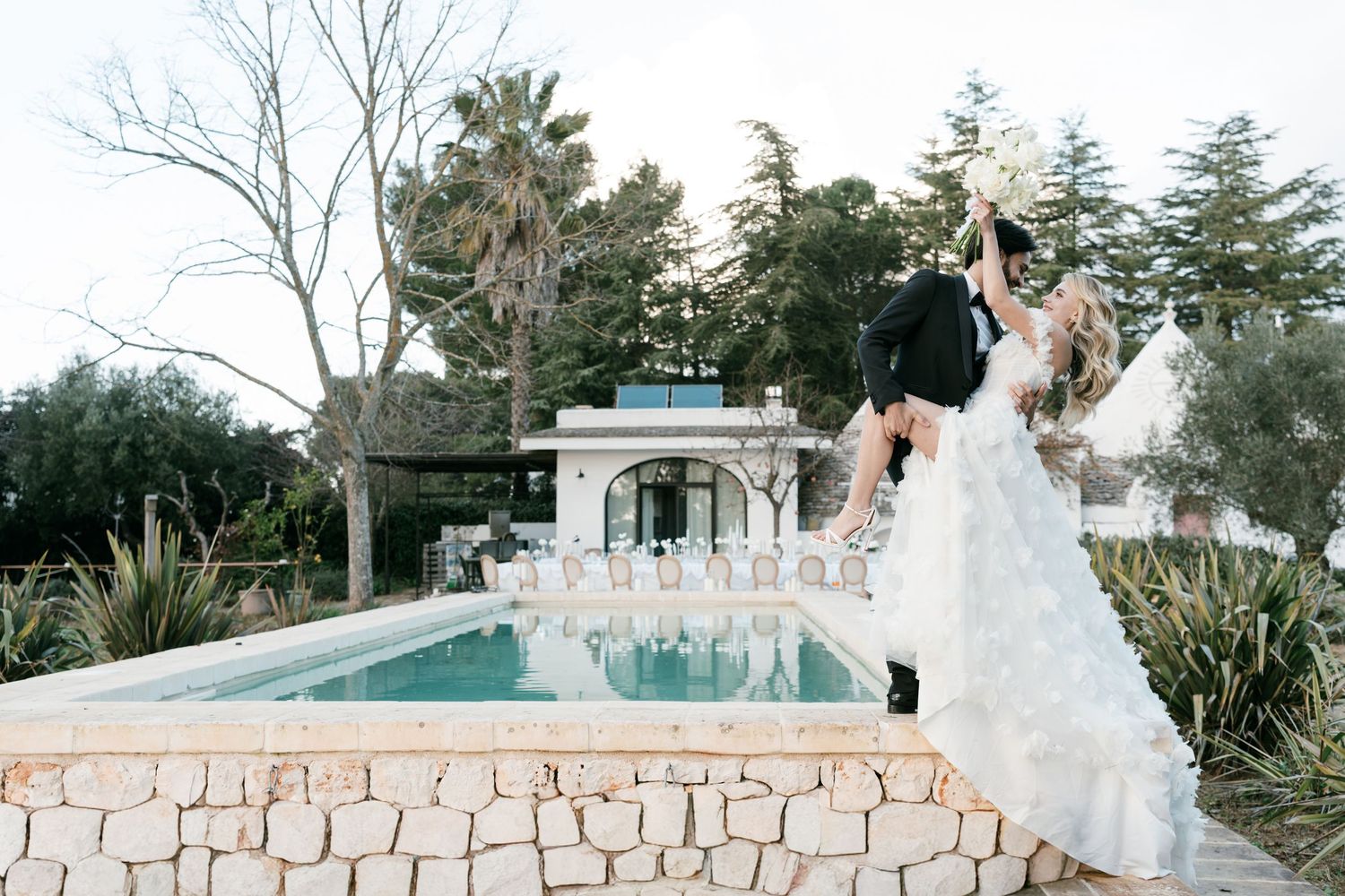 Bride in ruffled gown lifted by groom near elegant pool with palm trees at outdoor wedding venue.