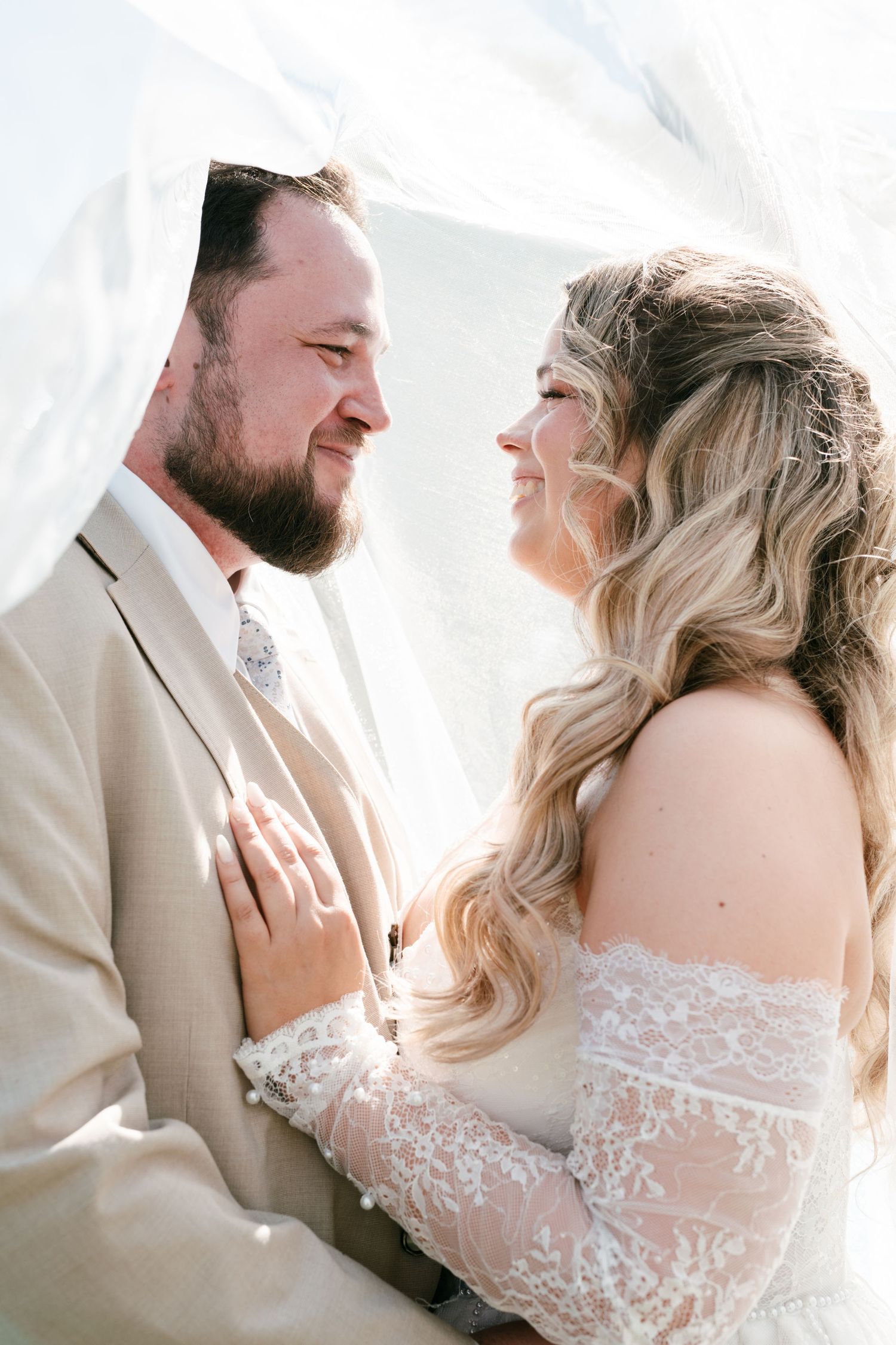 A bride and groom share a tender moment under a veil, smiling at each other in elegant wedding attire.