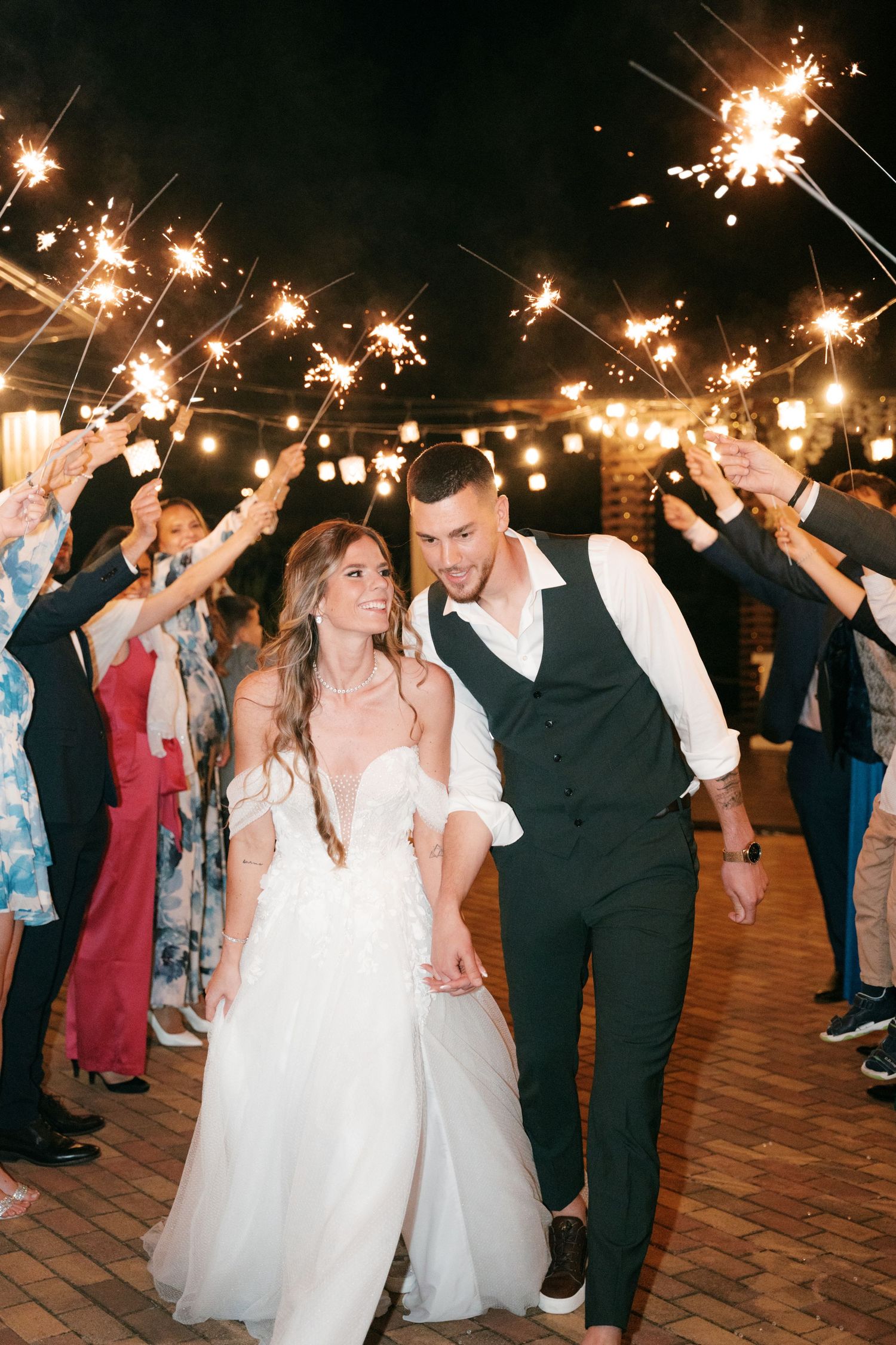 Bride and groom walk through sparkler tunnel surrounded by cheering wedding guests at night. Mallorca wedding photographer and filmmaker.