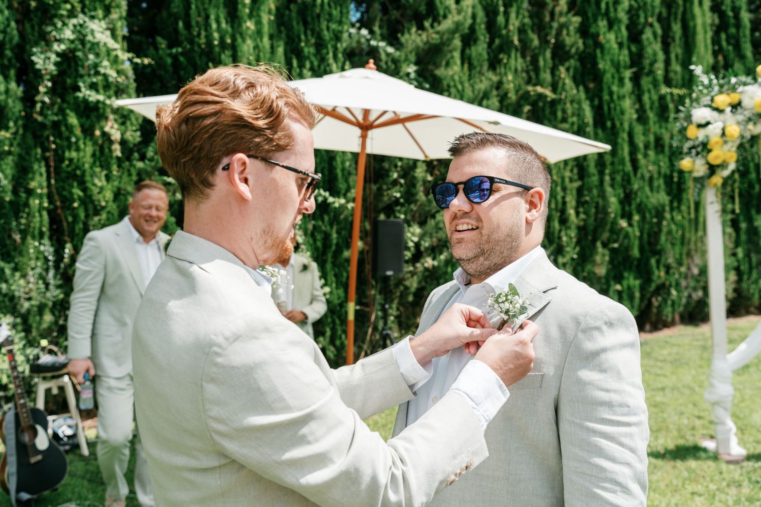Groomsman pinning boutonniere on groom in light suit at outdoor wedding ceremony with greenery backdrop. Mallorca wedding photographer