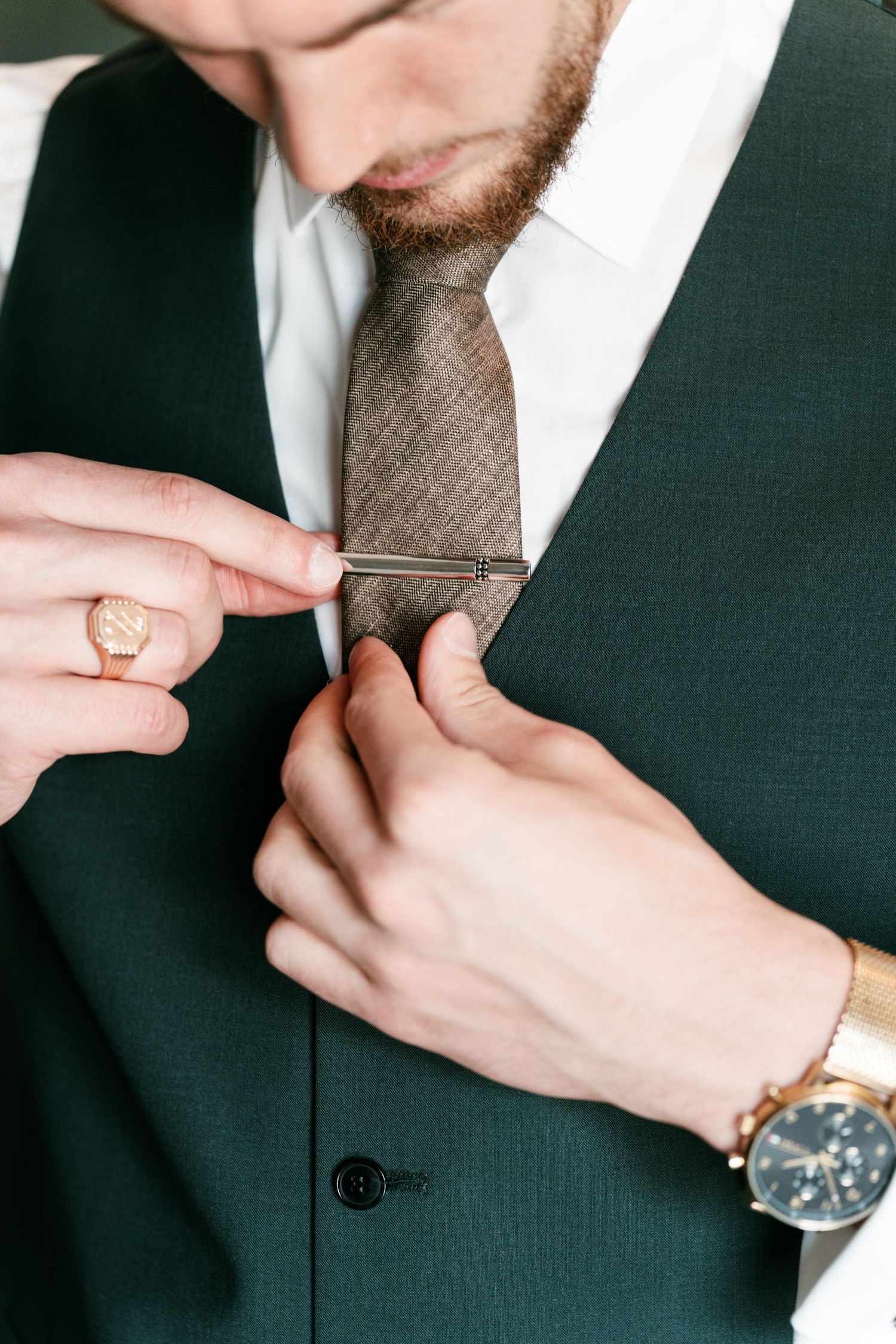 Groom in green suit adjusts tie clip on brown textured tie with gold watch. Mallorca wedding photographer and filmmaker.