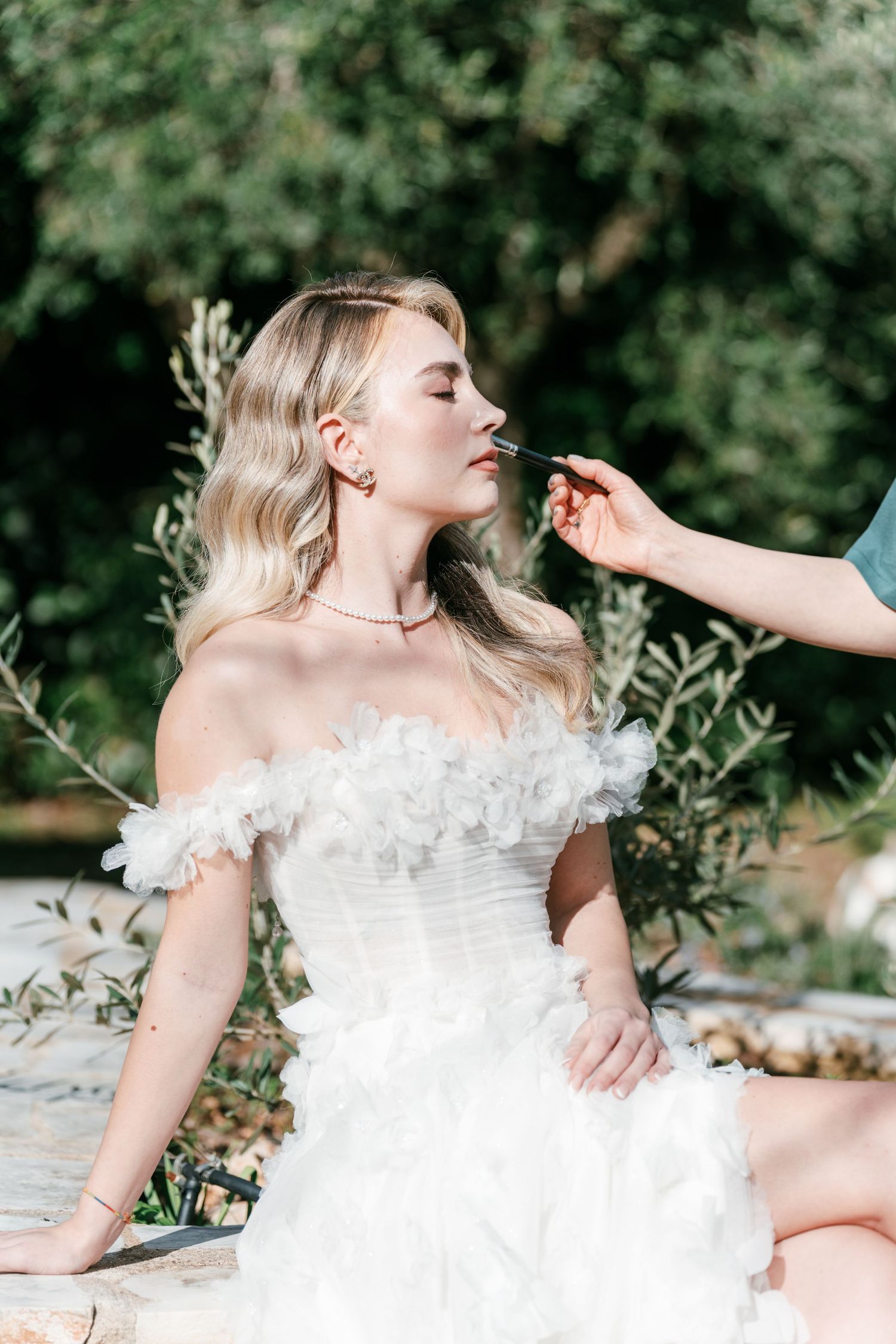 Bride in white off-shoulder ruffled gown gets lipstick applied by makeup artist outdoors. Puglia wedding photographer, film