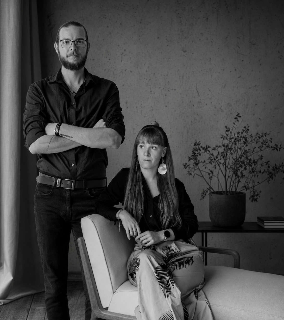 Black and white portrait of a man and woman in dark clothing, man standing with arms crossed, woman seated in armchair.