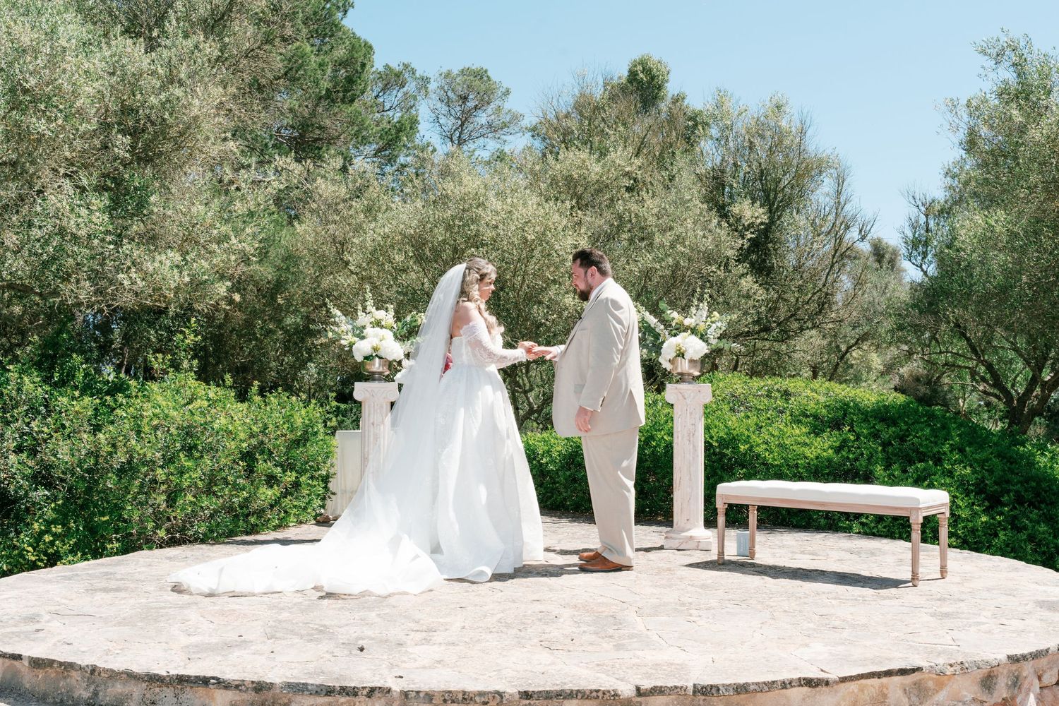 Bride and groom exchange vows outdoors at elegant garden wedding ceremony with white floral arrangements and lush greenery.