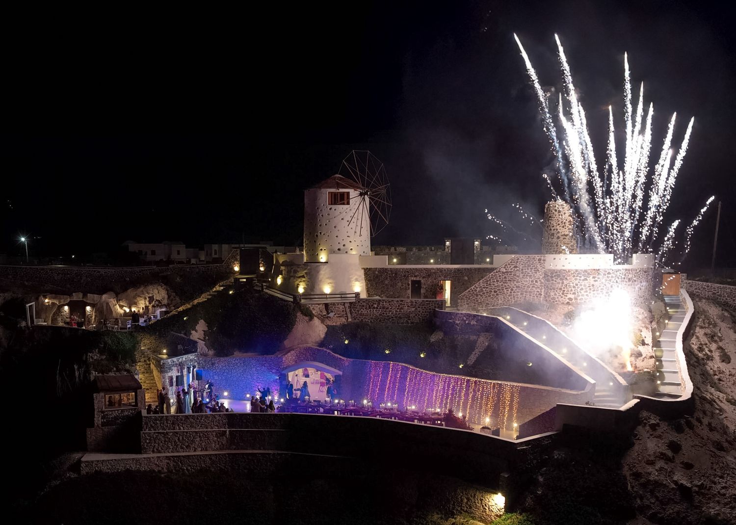 Fireworks illuminate a historic fortress at night during an outdoor concert event with colorful stage lighting.