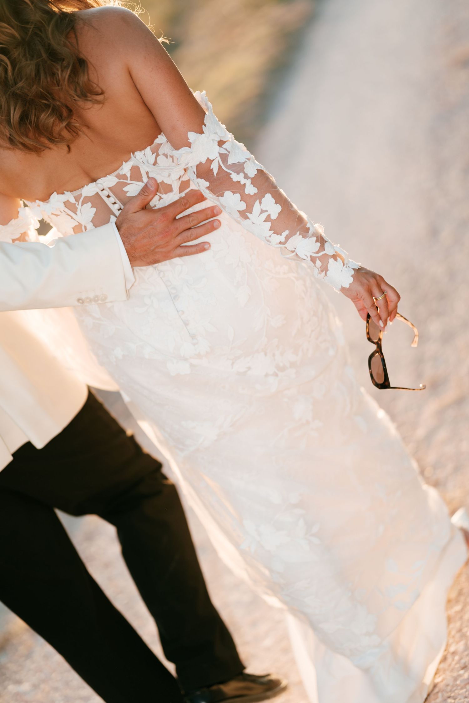 Bride in off-shoulder lace floral wedding gown held by groom outdoors in golden light.