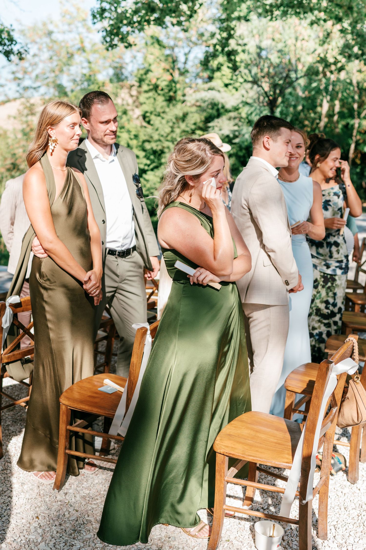 Emotional wedding guests in elegant green gowns and suits standing during an outdoor ceremony on a sunny day.