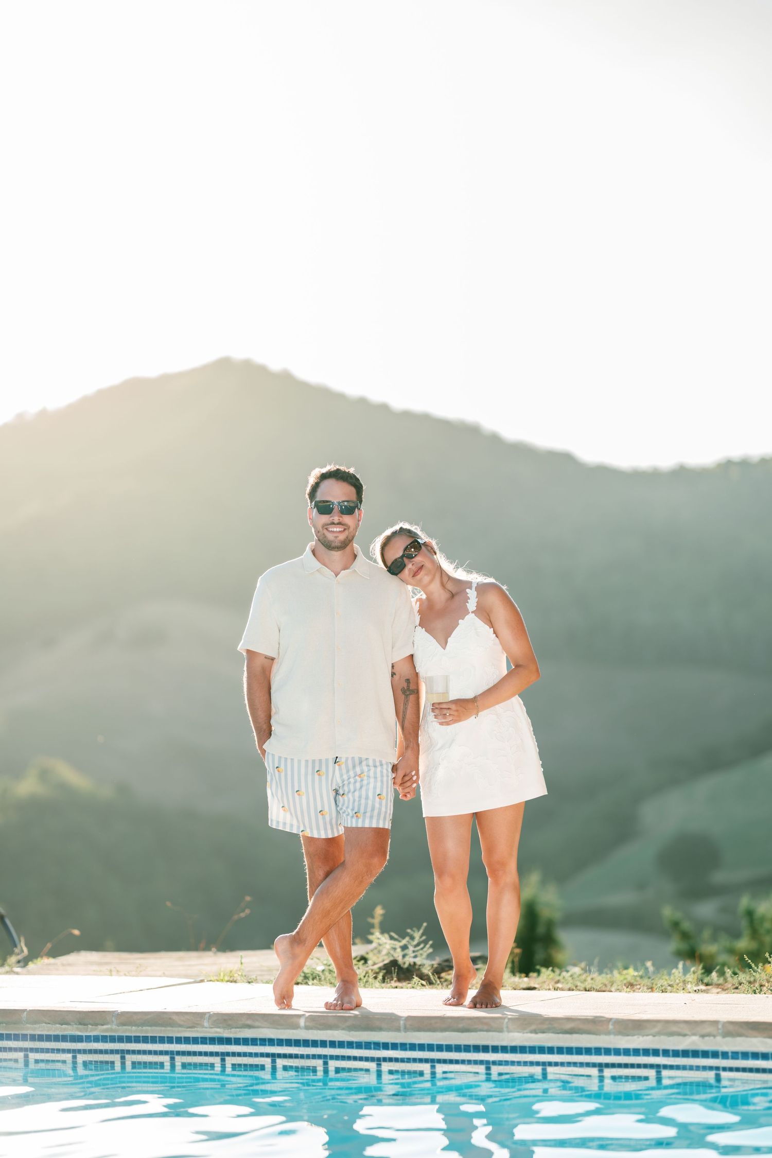 Couple in white outfits holding hands by a pool with green mountains in the background on a sunny day.