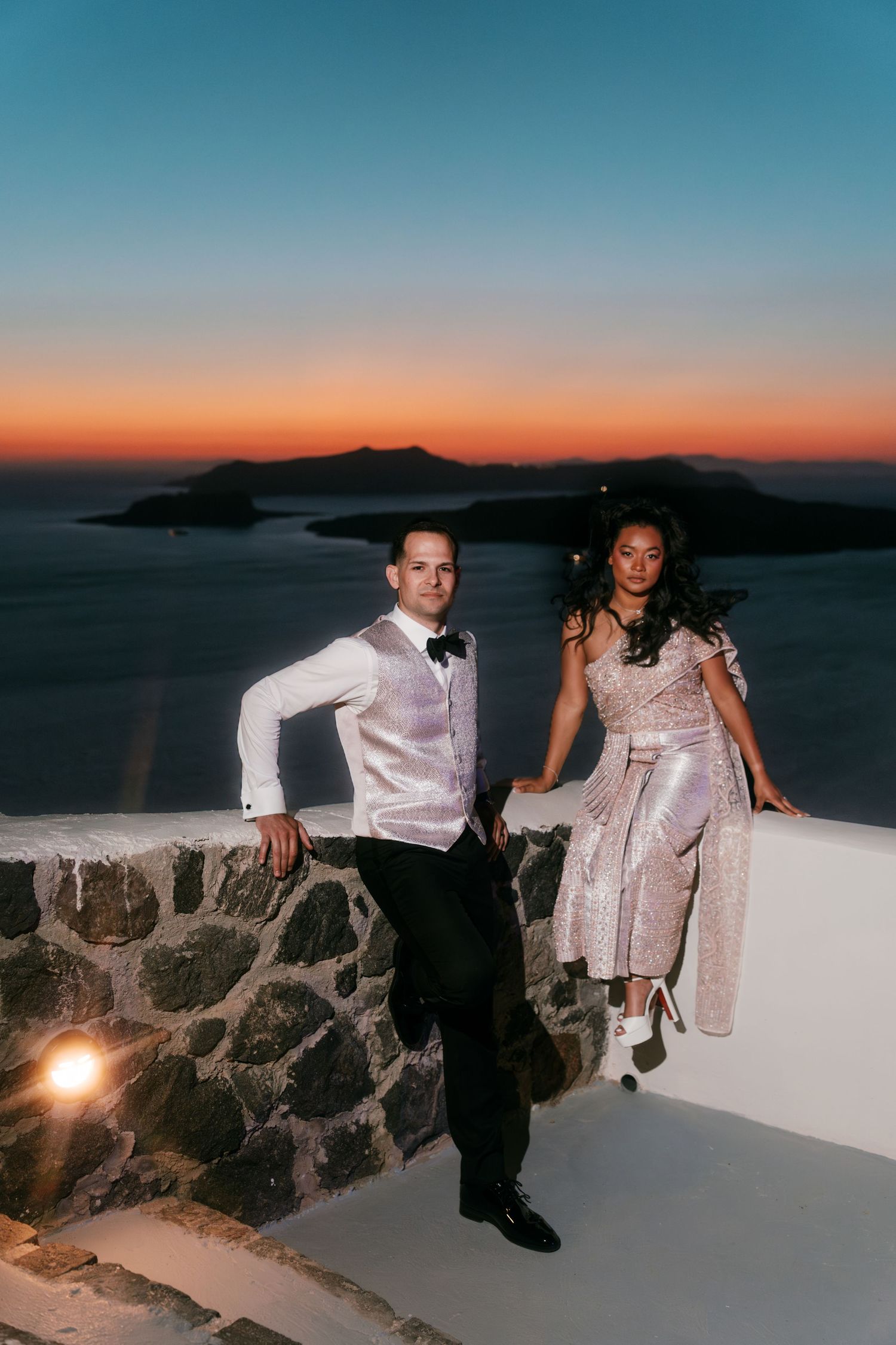 Couple poses romantically against a stone wall during a breathtaking Santorini sunset with caldera views.