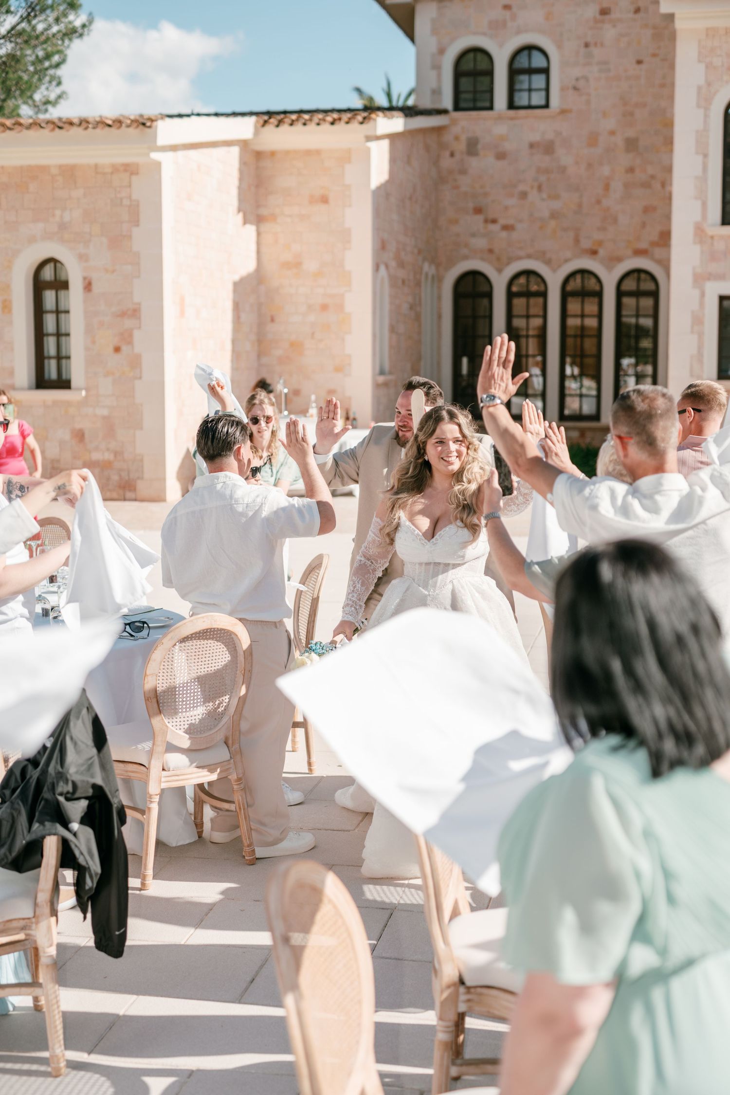 Bride and groom high-fiving guests at outdoor wedding reception at elegant stone venue on sunny day in Mallorca.