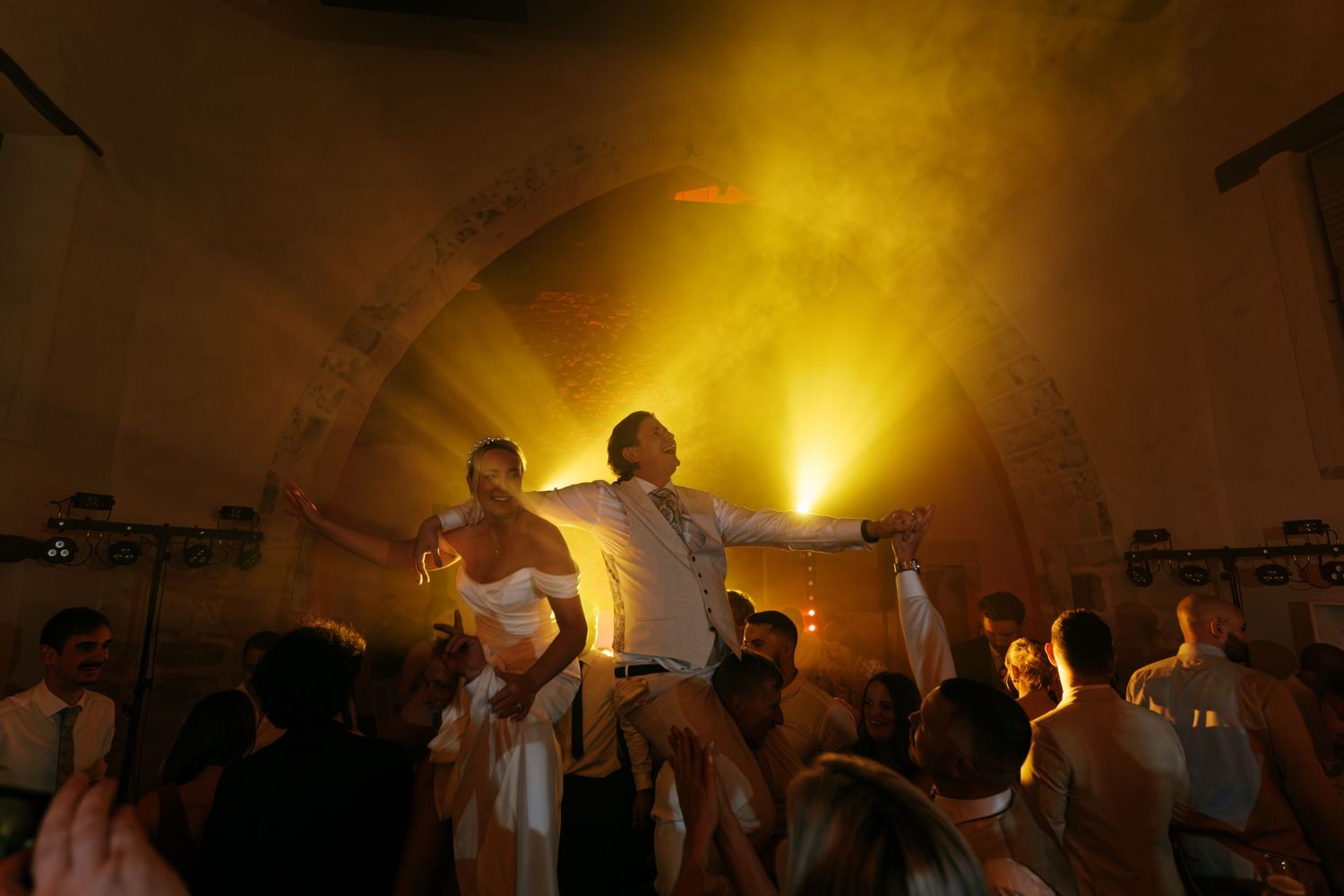 Wedding guests lift a bride and groom into the air on chairs during a lively reception dance under glowing yellow stage lights.