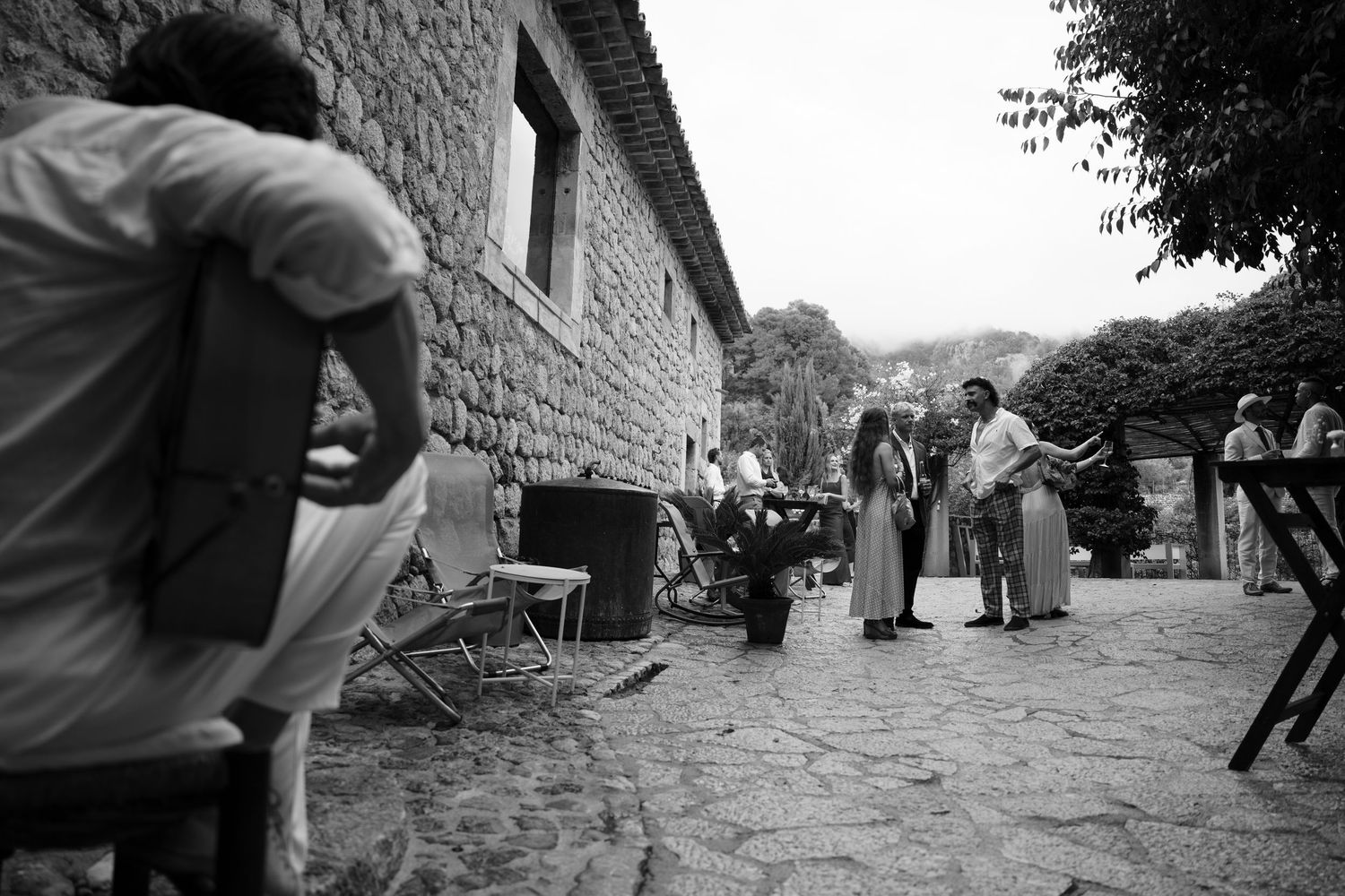 Black and white photo of wedding guests chatting on a stone patio beside a rustic stone building, with a seated man in the foreground.