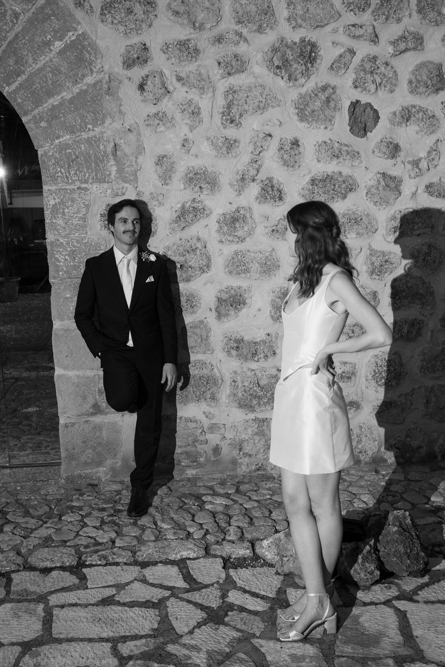 Black and white photo of a groom in a suit leaning against a stone arch while a bride in a short white dress watches on a cobblestone path.