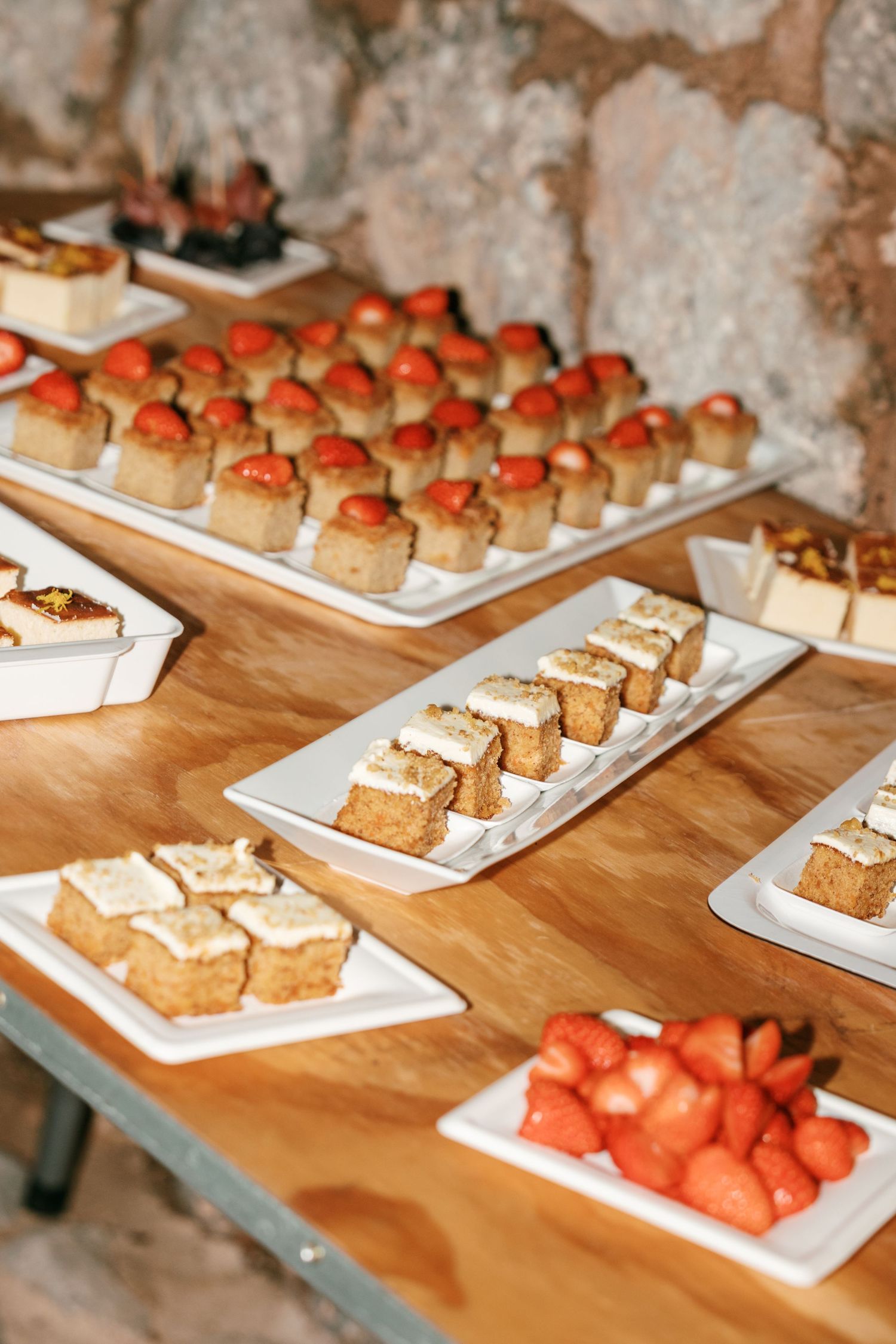 White platters of mini cake squares topped with fresh strawberries and cream on a rustic wooden table by a stone wall.